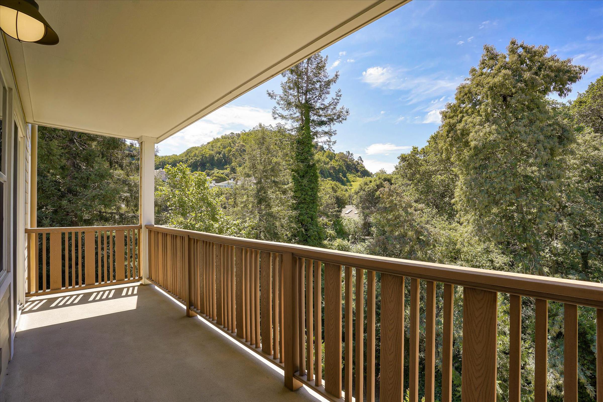 A spacious balcony with a wooden railing overlooking a lush green landscape, featuring tall trees and a clear blue sky with scattered clouds. The scene conveys a peaceful outdoor space ideal for relaxation and enjoying nature.
