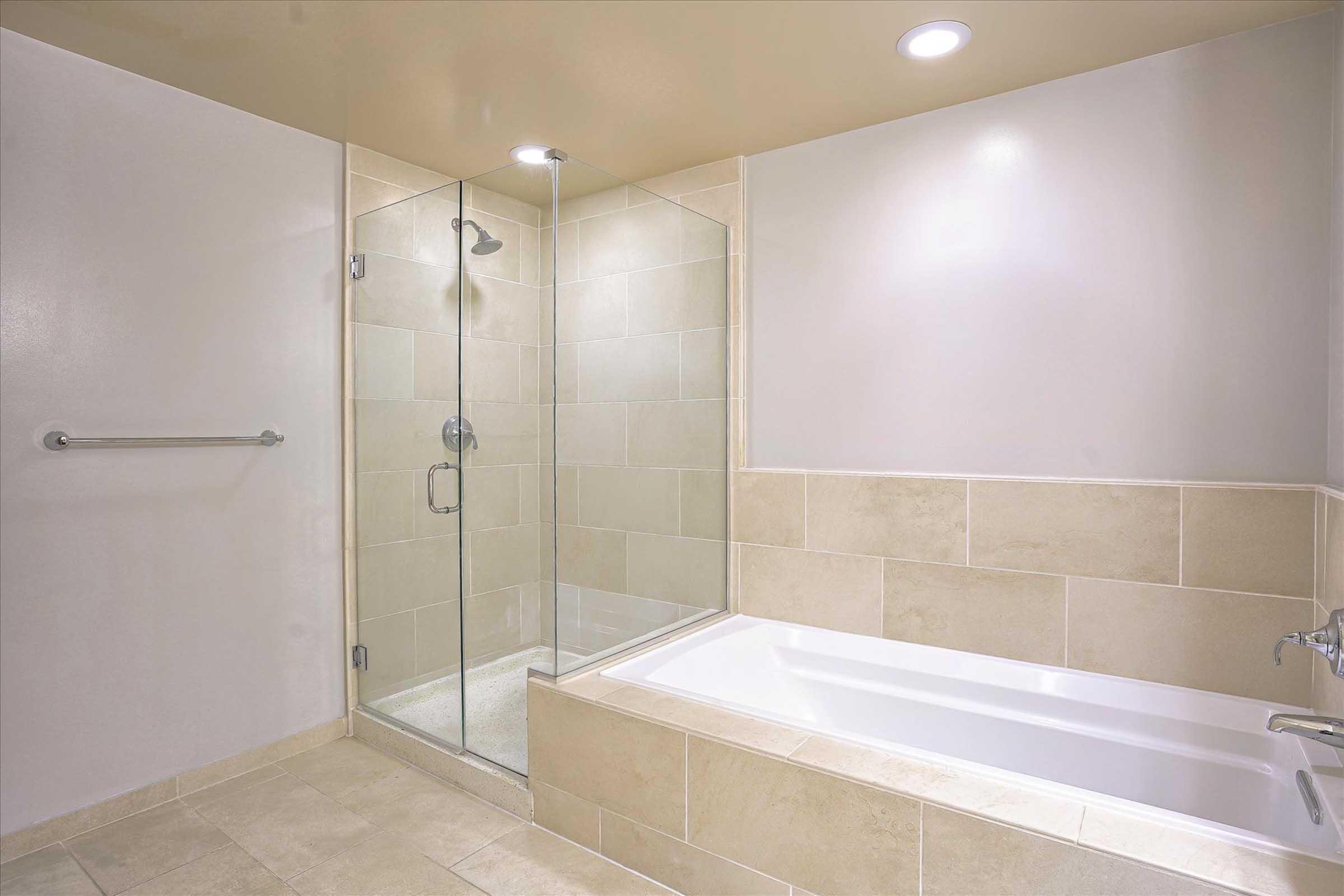 A modern bathroom featuring a glass-enclosed shower on one side and a built-in bathtub on the other. The floor and walls are tiled with light-colored stone, and there are recessed lights in the ceiling, giving the space a bright and clean appearance.