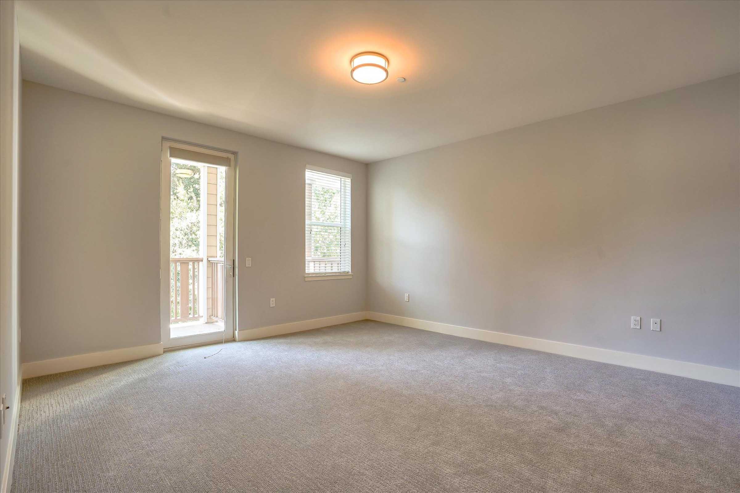 A spacious, empty room with light gray walls and a carpeted floor. Natural light streams in through a window and a glass door that leads to a small balcony. The ceiling features a simple light fixture, creating a bright and inviting atmosphere.