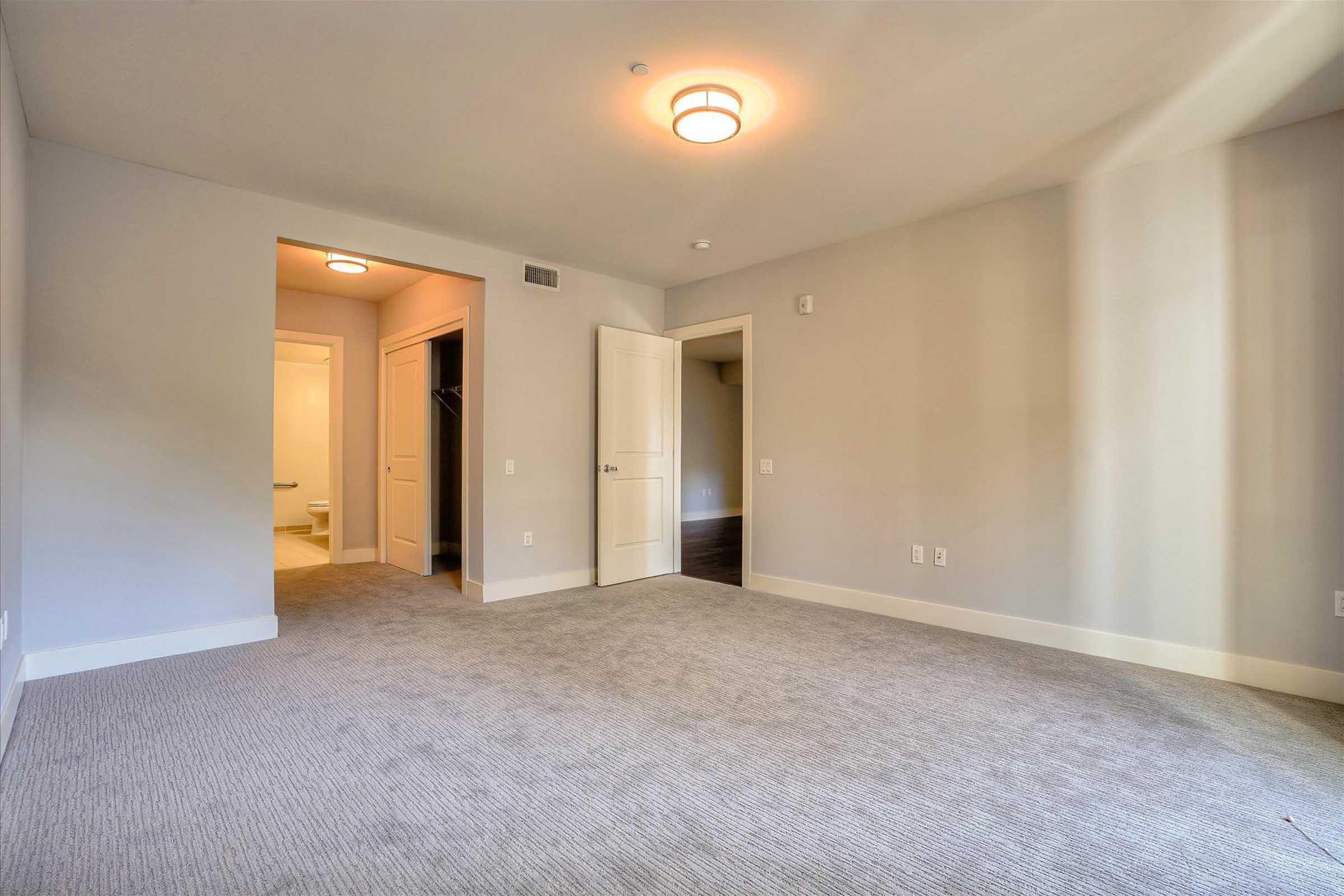 Spacious, empty room featuring light gray walls and carpet. A doorway leads to a small hallway with a bathroom visible at the end. Light fixtures on the ceiling provide illumination, and natural light enters from the visible doorway. The overall ambiance is bright and modern.