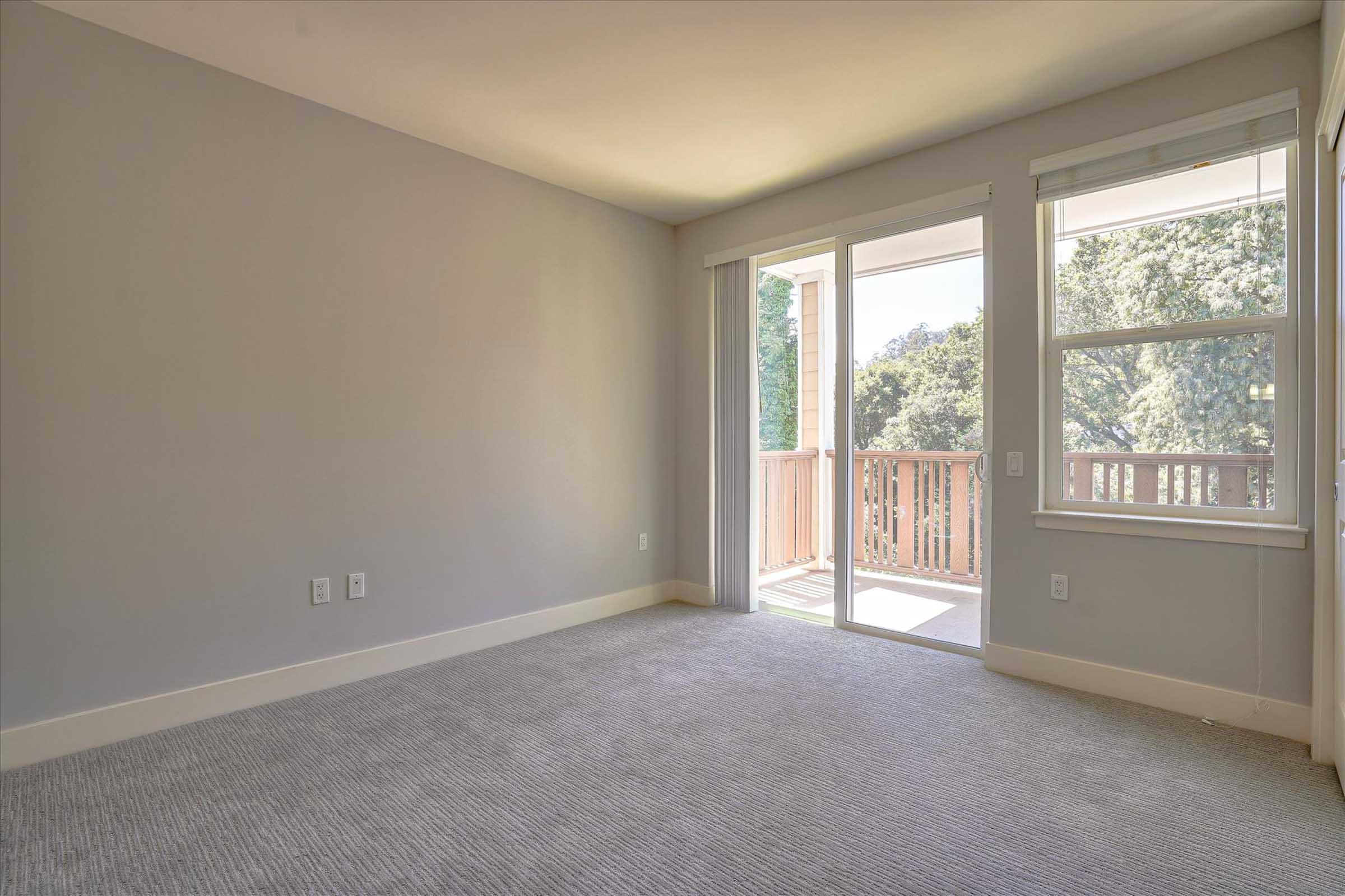 A spacious, unfurnished room with light gray walls and carpet flooring. It features a large window with natural light coming in, overlooking a balcony. The atmosphere is bright and inviting, suitable for various interior designs.