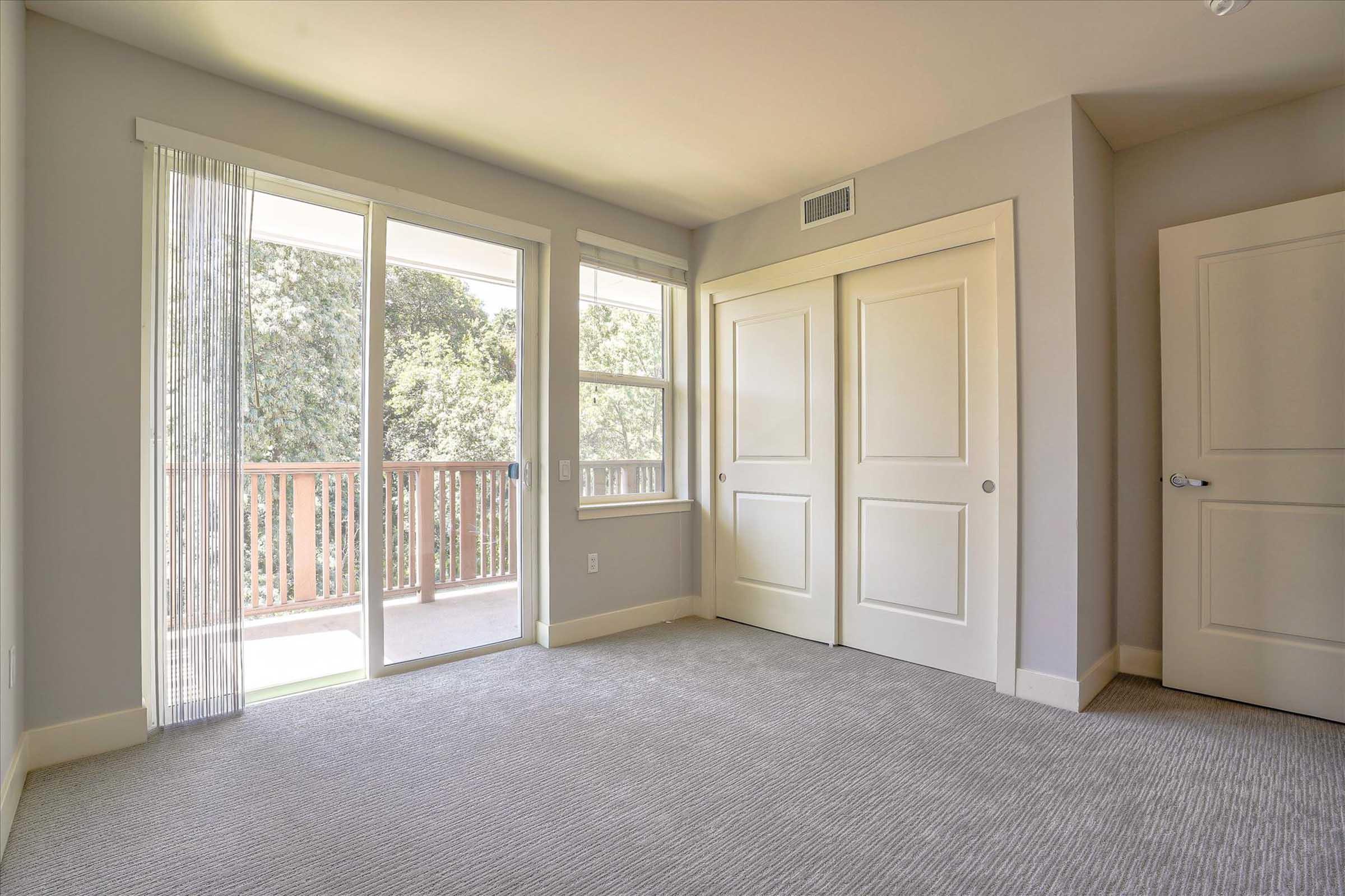 A bright, empty room with light gray walls and carpet. It features a large window with sheer curtains letting in natural light, a door leading to a balcony, and two white closet doors against the far wall. The overall aesthetic is clean and modern.