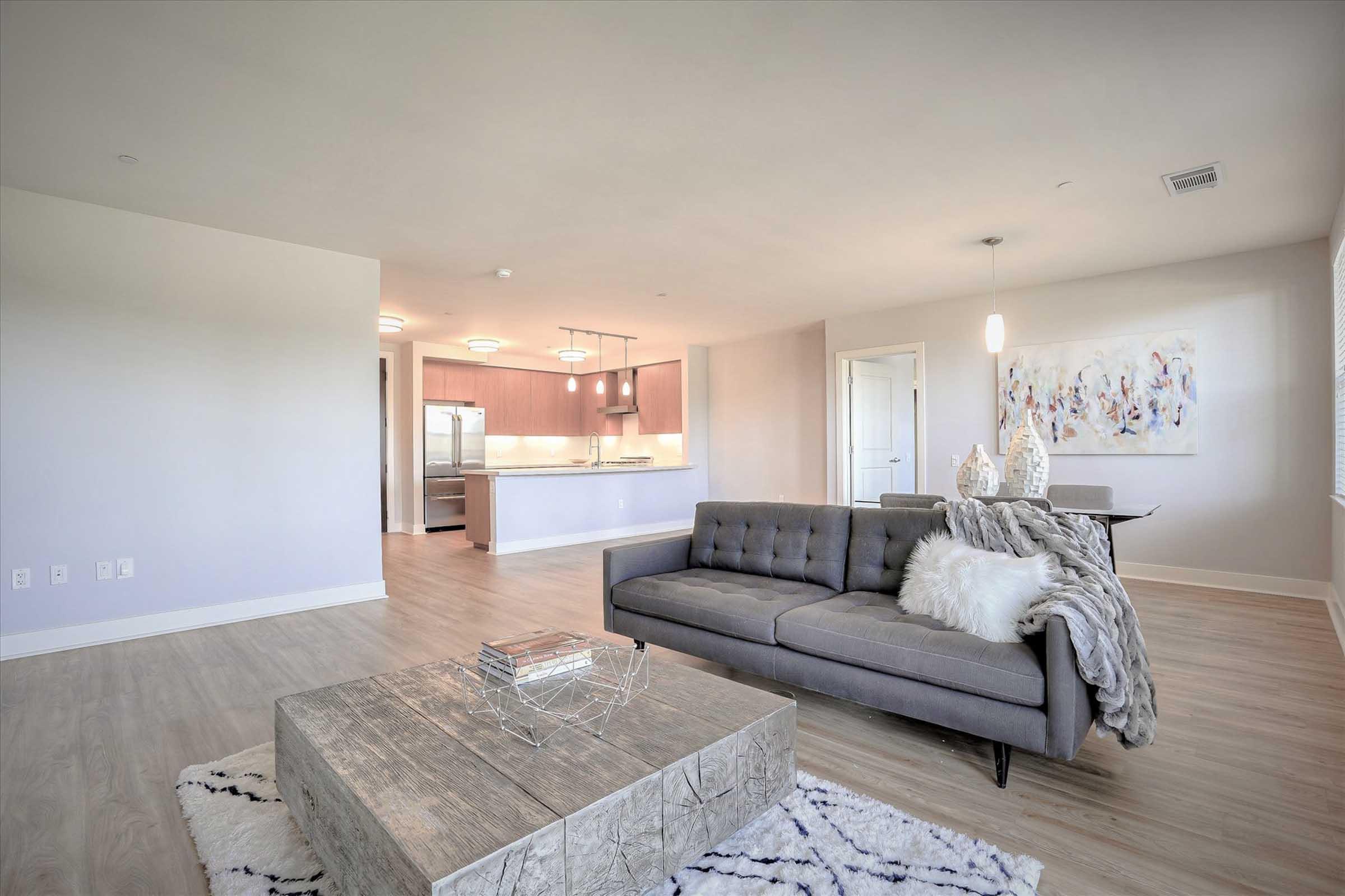 A modern living room featuring a gray sofa with cushions, a textured white throw, and a wooden coffee table. The room has light wood flooring, large windows for natural light, and a glimpse of an open kitchen area with sleek appliances and cabinetry. Wall art decorates the space, adding a contemporary touch.