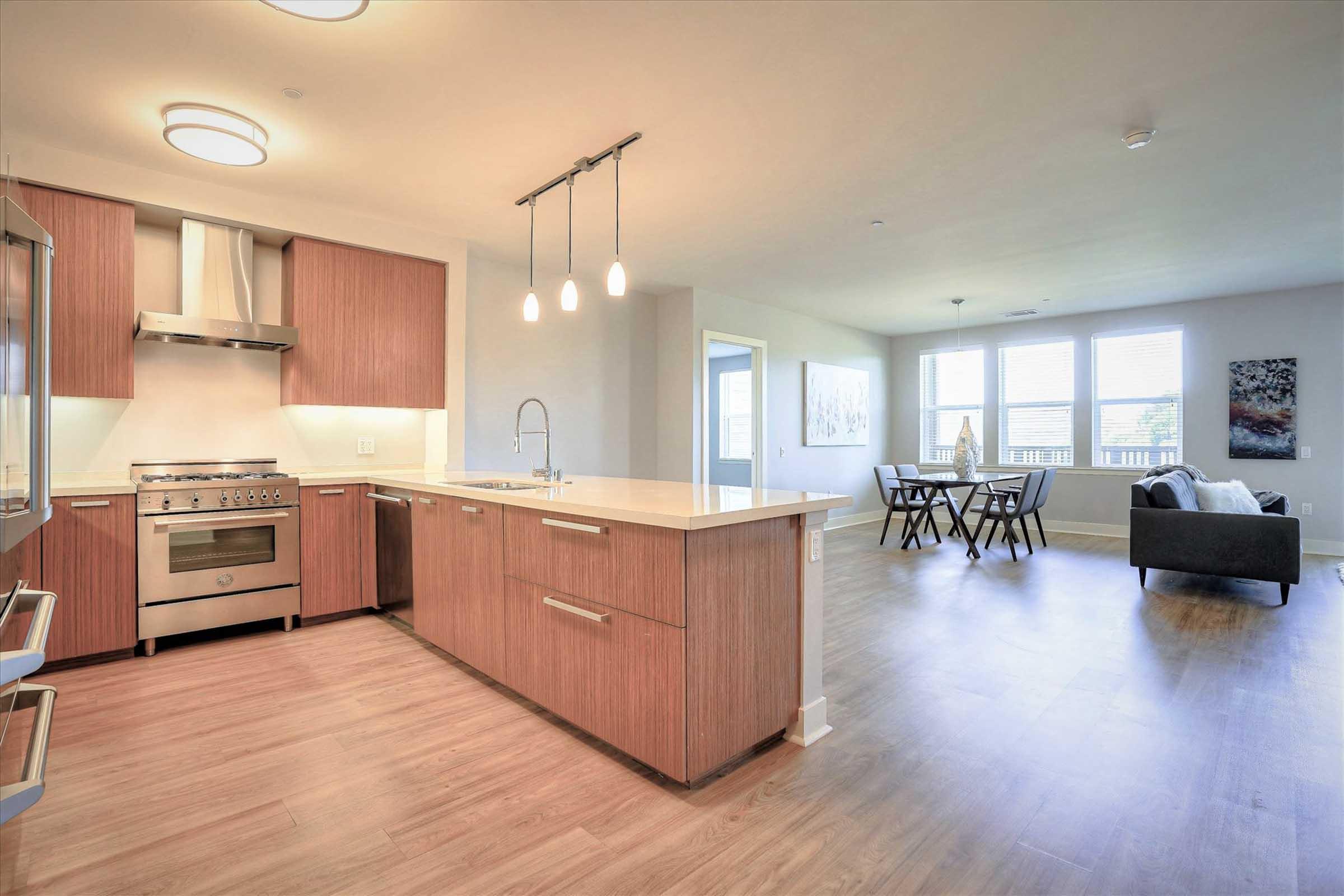 A modern kitchen featuring wood cabinets, a stainless steel oven, and a sink with a countertop. To the right, an open living area with a dining table and chairs, large windows, and stylish artwork on the walls. The floor is a warm wood finish, creating an inviting atmosphere.
