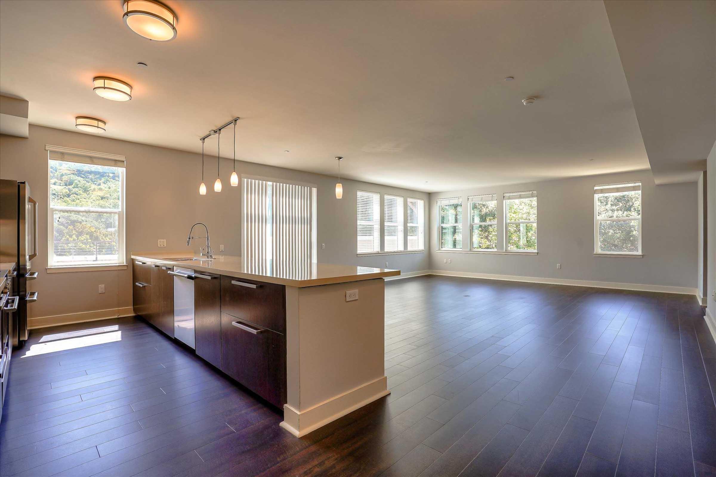 A spacious modern living area featuring large windows that provide natural light, hardwood flooring, and a kitchen with dark cabinetry and pendant lighting. The kitchen is open to the living space, creating an airy atmosphere, with a neutral color palette and a serene view outside.