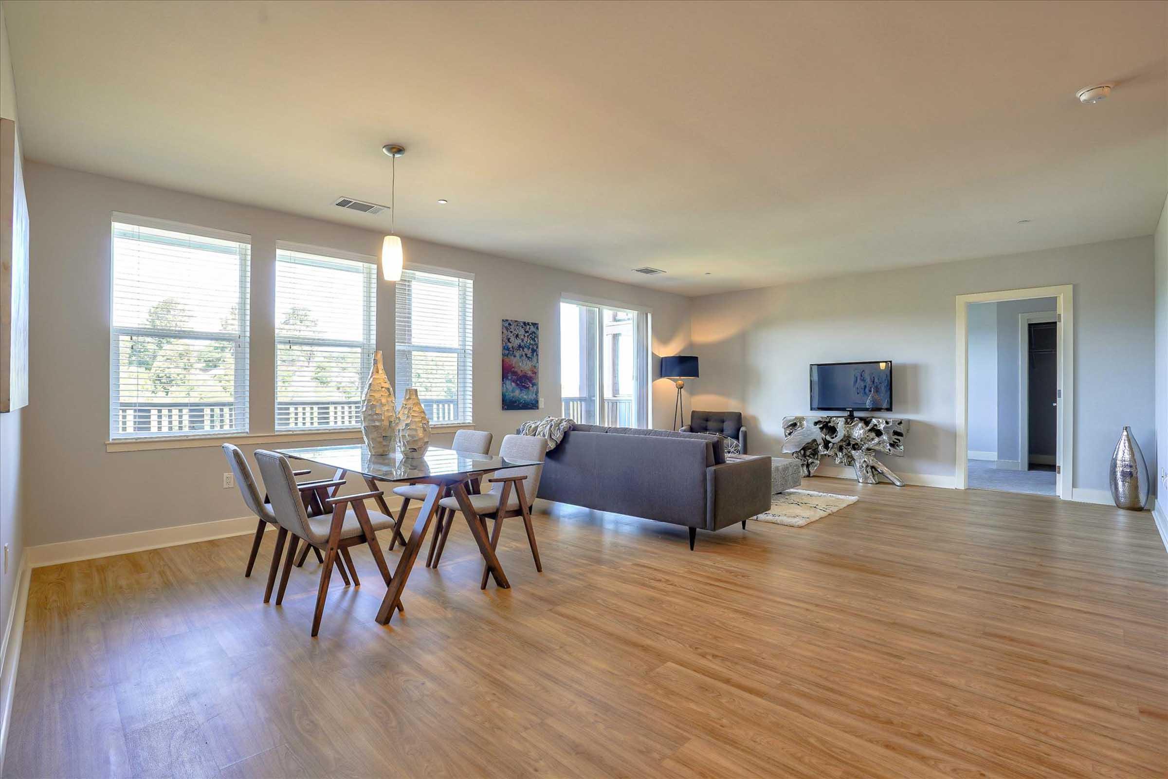 Bright and spacious living area featuring a modern gray sofa, wooden dining table with chairs, and a stylish TV stand. Large windows let in natural light, and there are decorative elements like a sculpture and a plant. The flooring is wooden, adding warmth to the contemporary design.