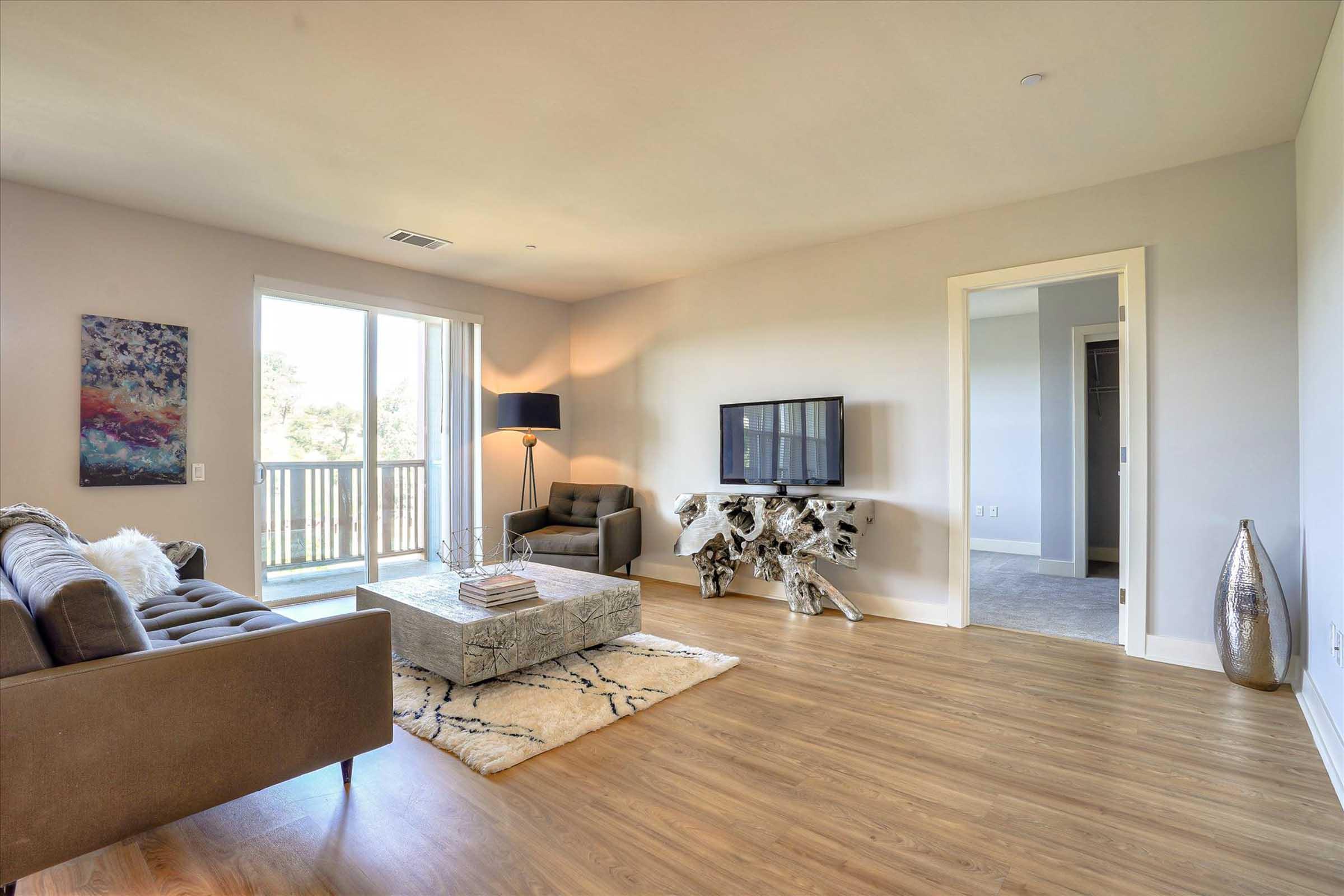 A modern living room featuring a brown sofa, a glass coffee table, a flat-screen TV on a decorative stand, and a cozy chair. Large windows allow natural light, revealing a balcony. The floors are wood, and there are stylish decor elements including a vase and wall art.