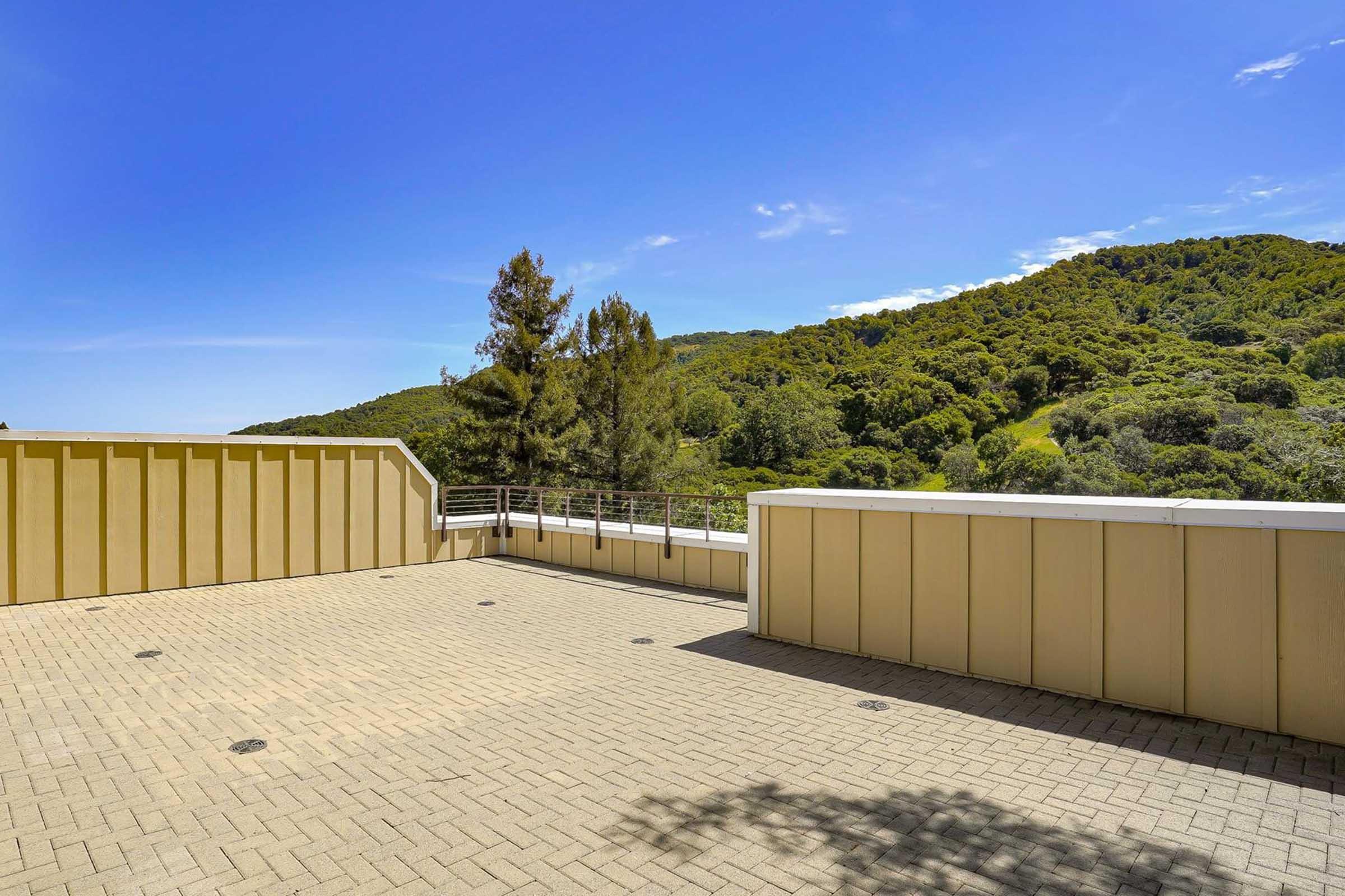 A spacious outdoor patio with a brick floor, overlooking a lush green hillside under a clear blue sky. The area features low walls and trees in the background, creating a serene and inviting atmosphere, ideal for relaxation or gatherings.