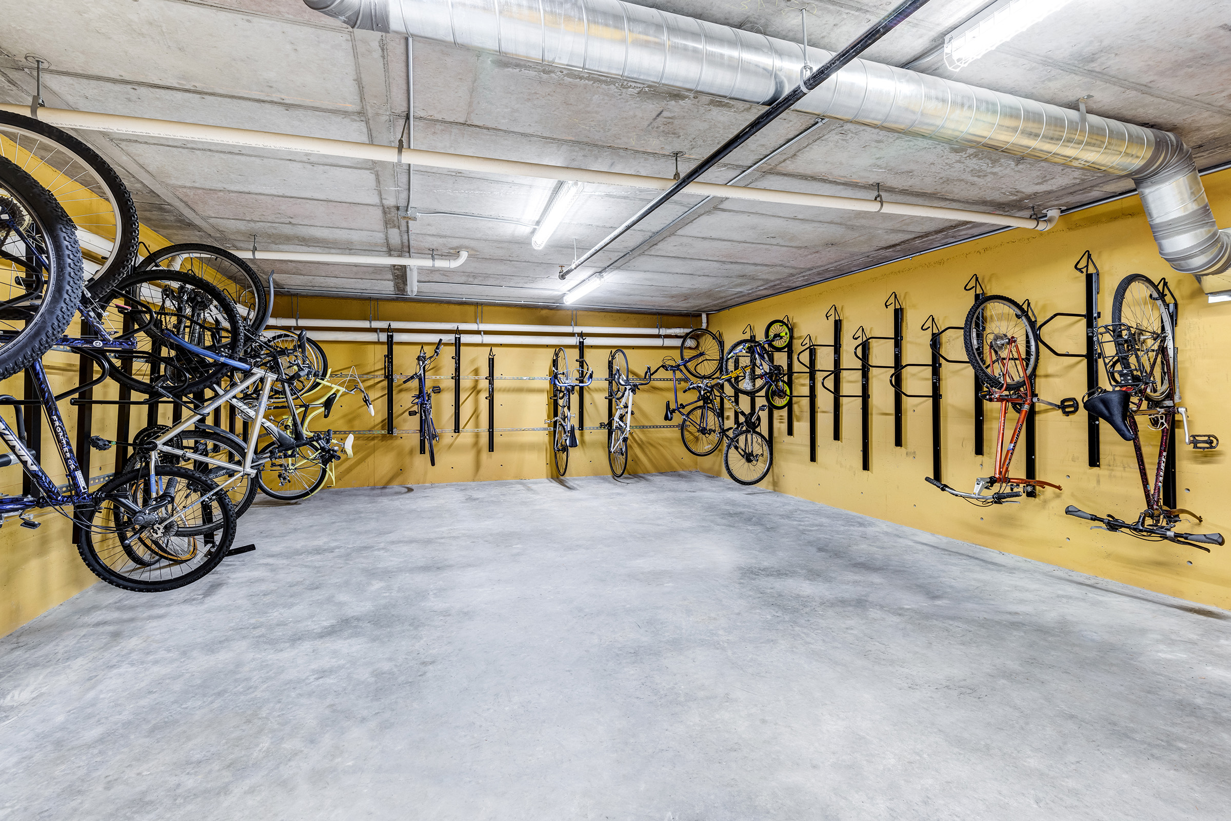 A spacious indoor bike storage area with several bicycles mounted on wall racks. The walls are painted yellow, and the floor is gray concrete. Overhead, there are exposed pipes and bright overhead lighting illuminating the space.