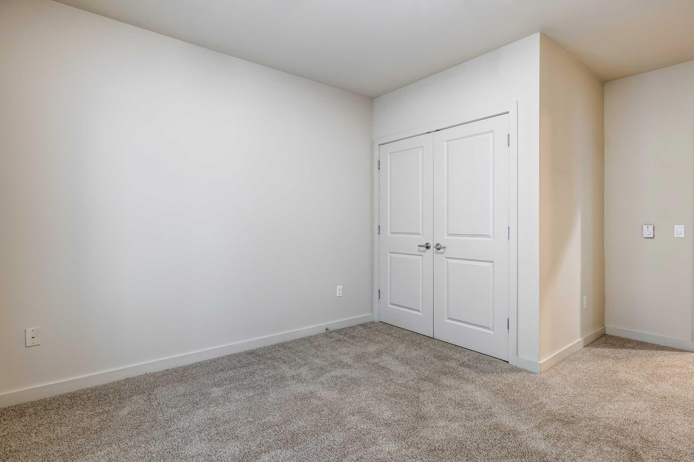 A spacious, empty room with light beige walls and a plush, beige carpet. On one side, there are white double doors leading to a closet or another space. The absence of furniture gives the room a clean, open feel, ideal for personalization.