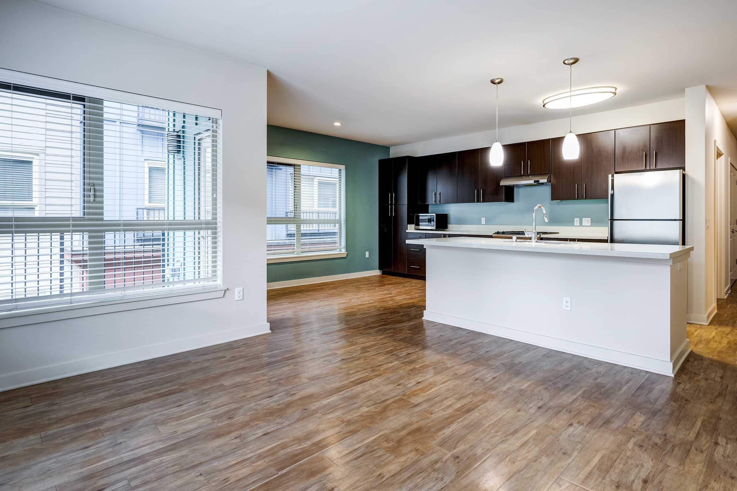 Modern open-concept kitchen and living area featuring hardwood floors, white cabinetry, and a large island. The kitchen includes stainless steel appliances and pendant lighting. Large windows provide natural light, with a green accent wall adding a pop of color.