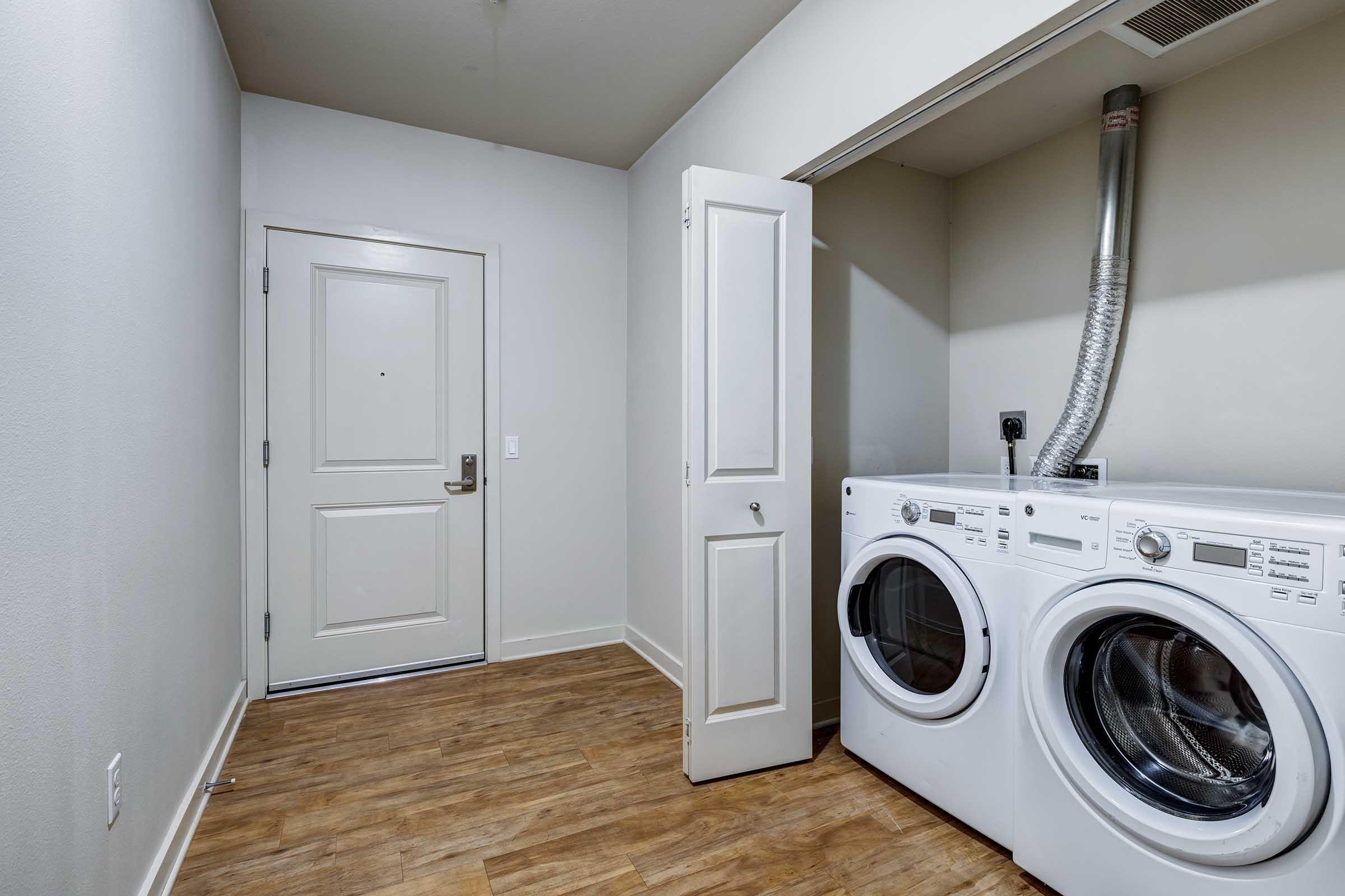 A clean laundry room featuring a white washer and dryer stacked side by side. There's a door leading outside and a small closet space to the left. The walls are a light color, and the floor is a wood-like laminate. The overall space is bright and uncluttered.
