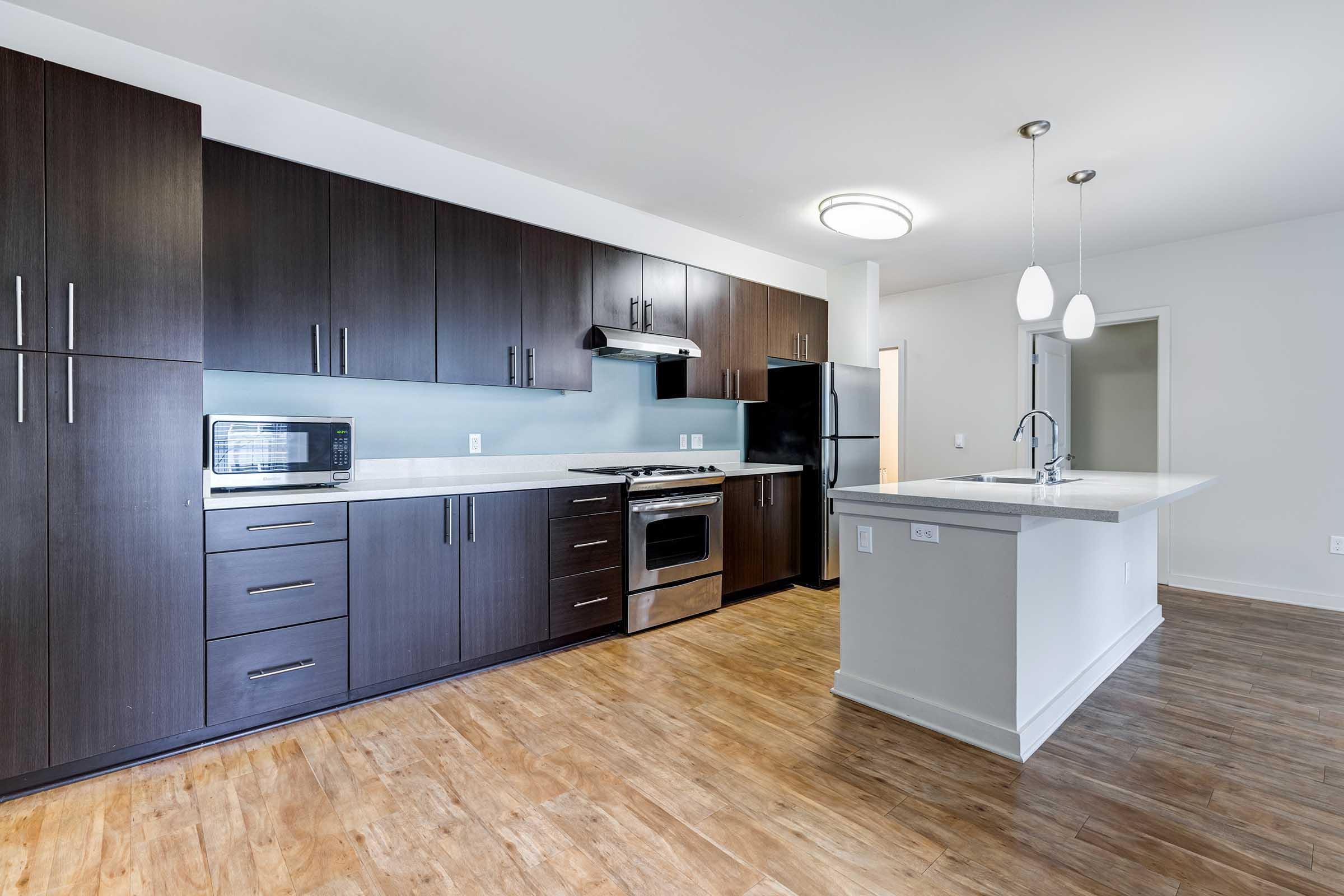 Modern kitchen with dark wood cabinetry, stainless steel appliances including a microwave and oven, and a central island with a sink. The flooring is light wood, complementing the sleek design. Bright lighting fixtures hang above the countertop, and the space is open and airy.