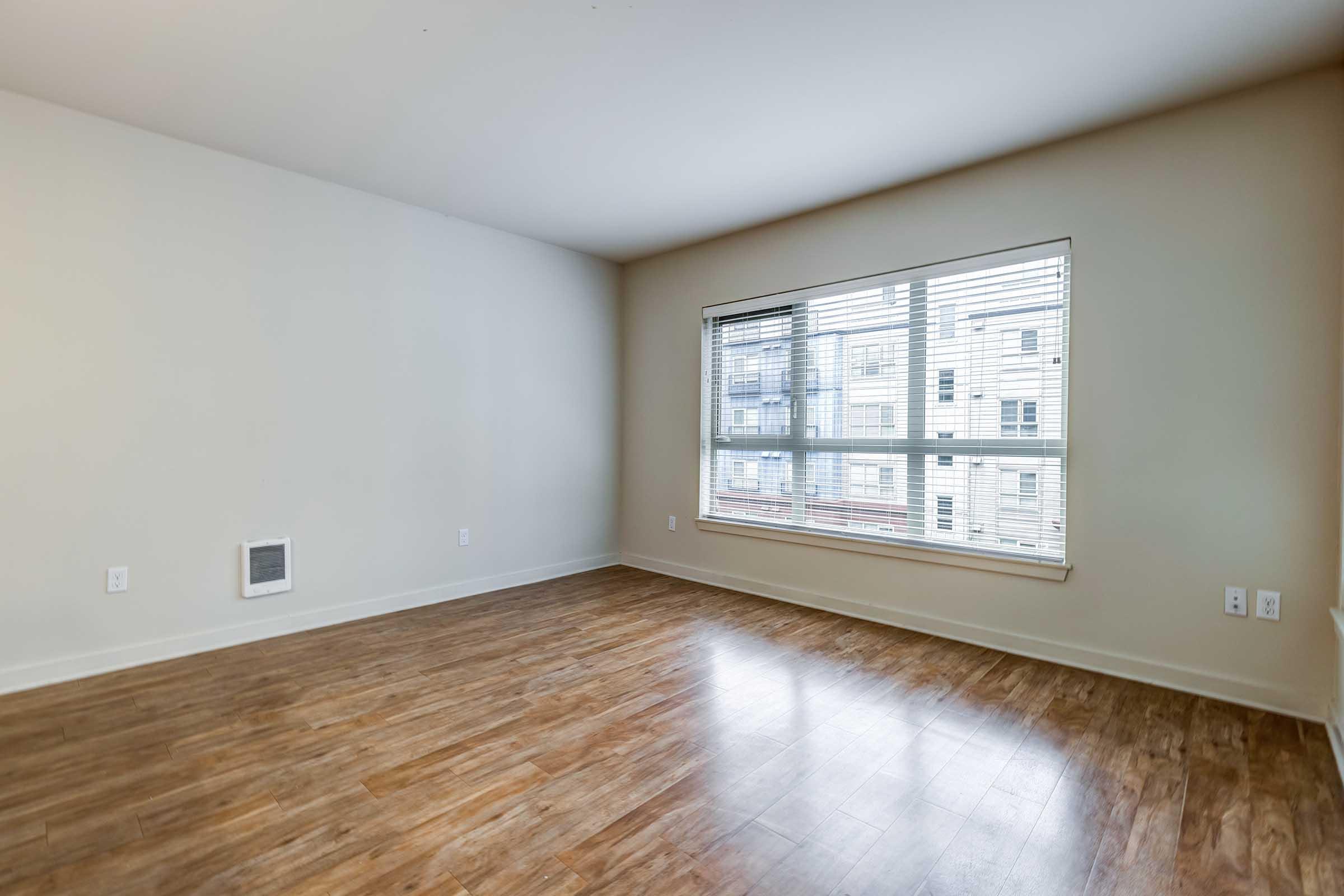 A spacious, empty room with light-colored walls and a large window featuring horizontal blinds. The floor is made of laminate wood, and there is an air vent on one wall. Natural light fills the space, creating a bright and inviting atmosphere.