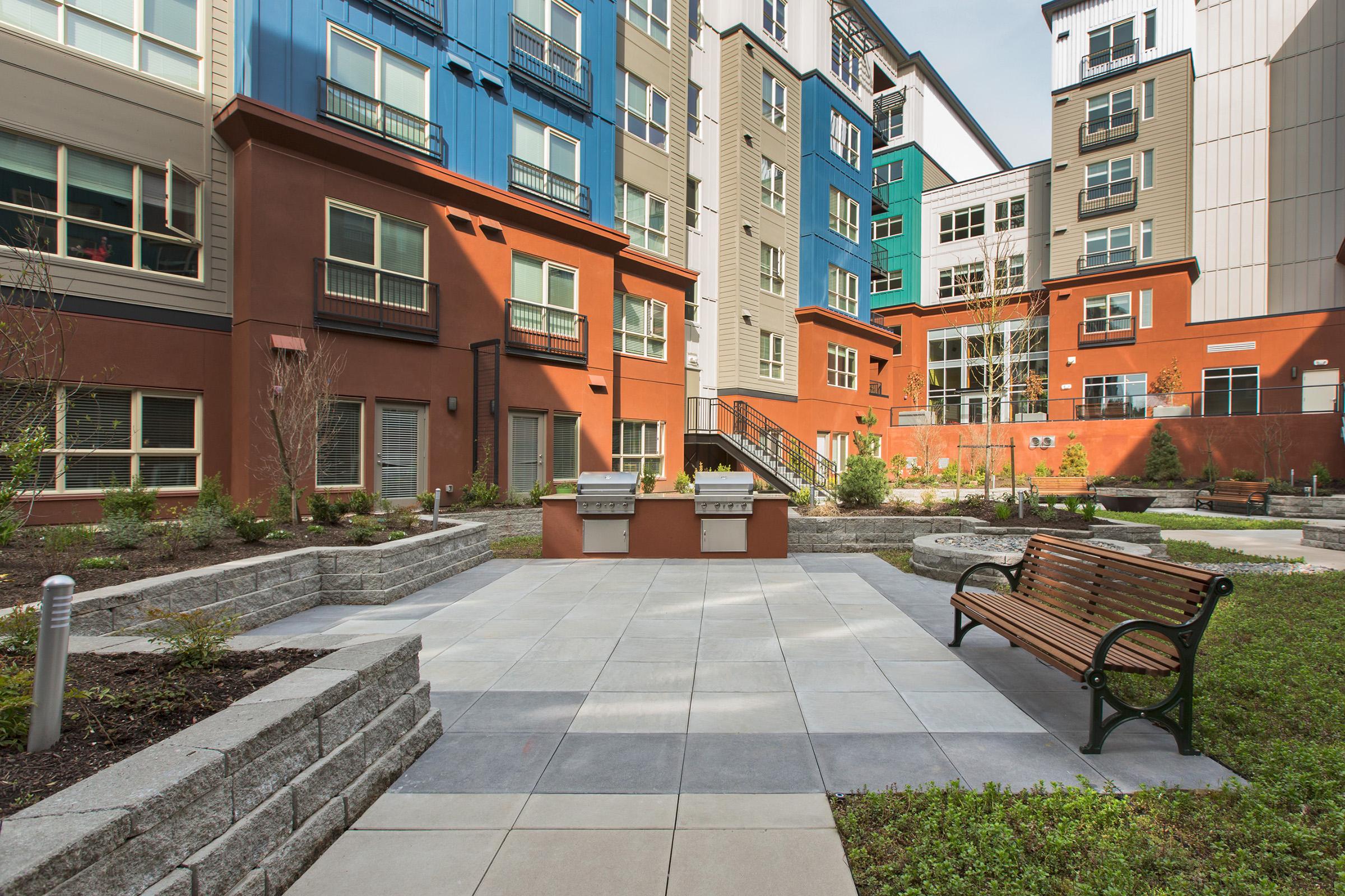 A modern urban courtyard featuring a paved sitting area with a wooden bench, surrounded by landscaped gardens.