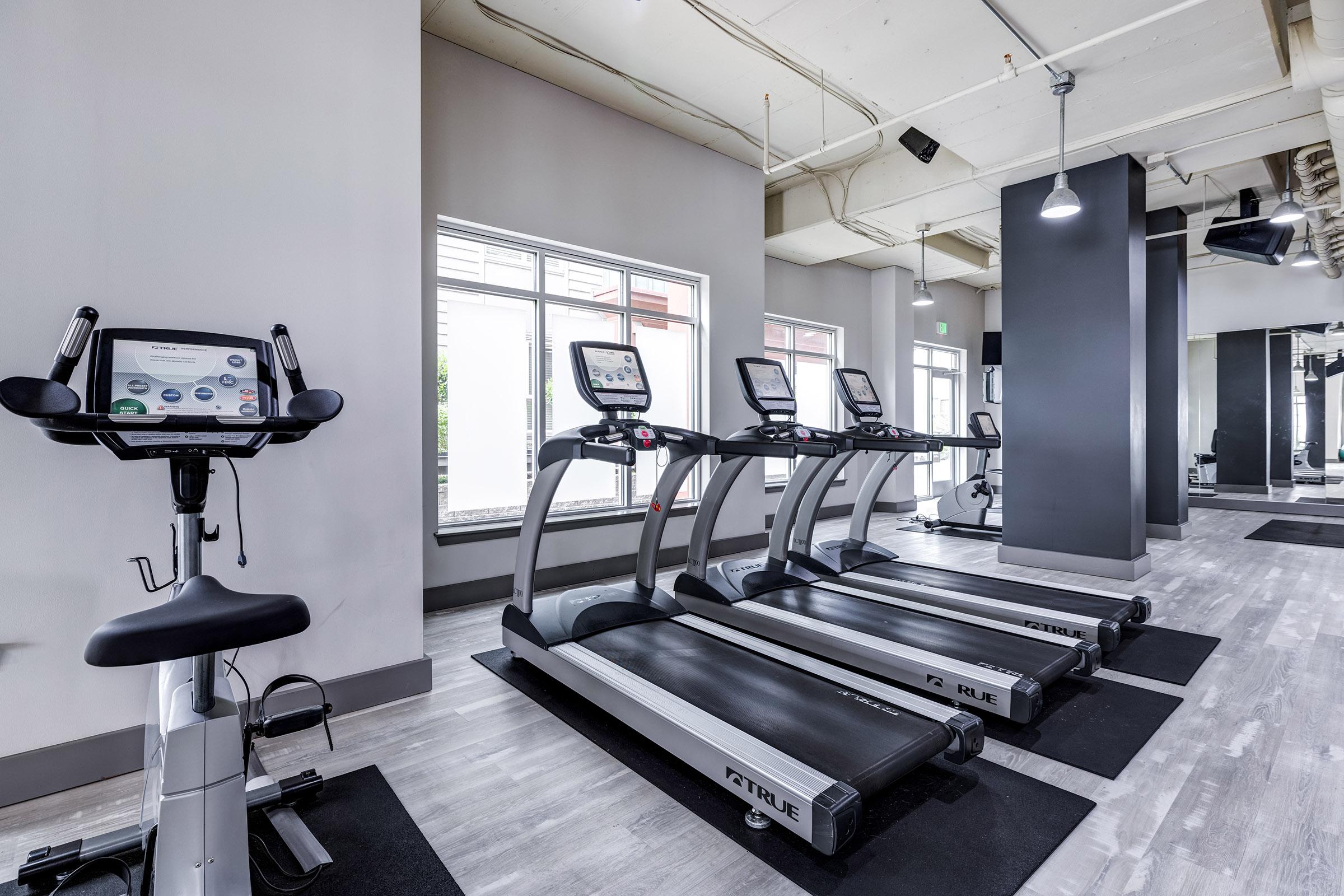A modern gym interior featuring several treadmills lined against a wall with large windows, providing natural light. A stationary bike is positioned nearby on a rubber mat.