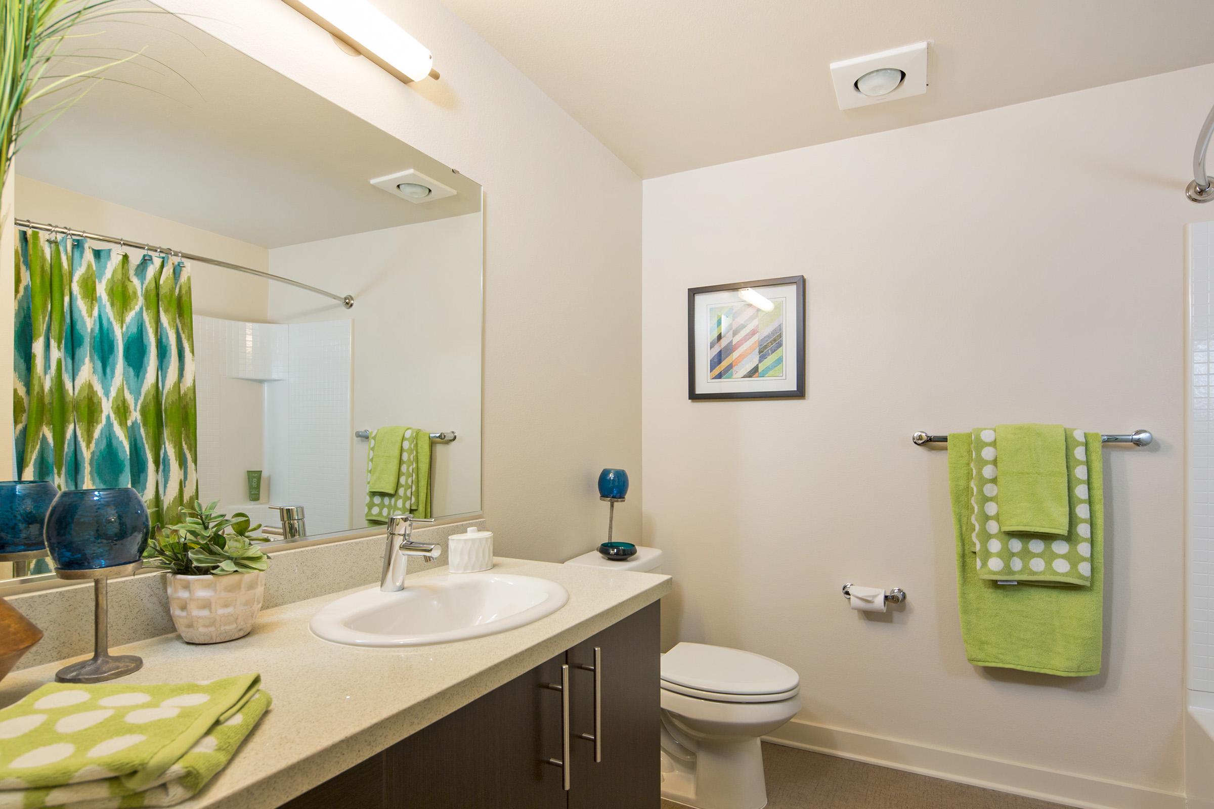 A modern bathroom featuring a white countertop with a sink, a mirror above it, and a colorful shower curtain with green and blue patterns.
