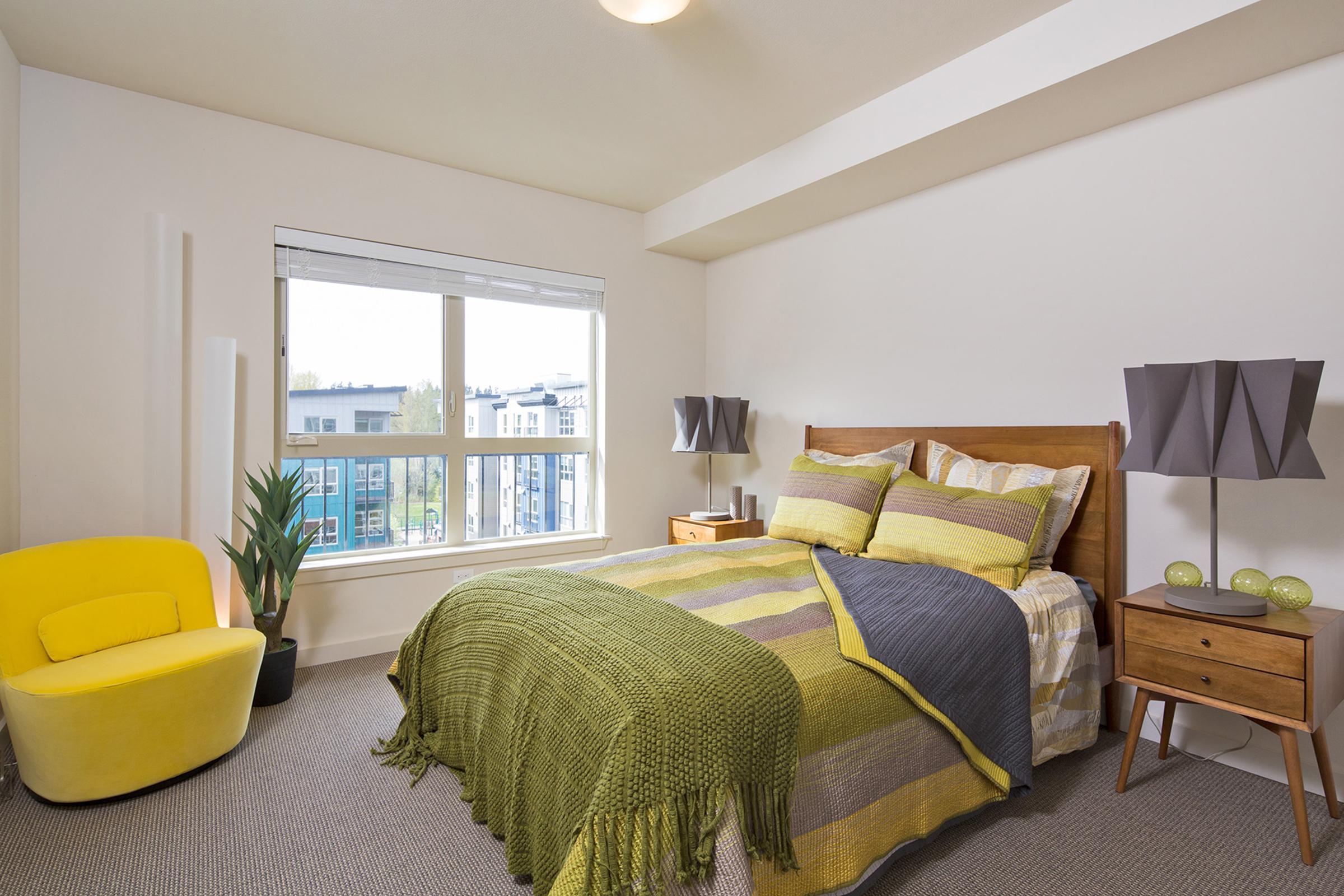 A modern bedroom featuring a queen-sized bed with green and gray bedding, a yellow accent chair, and a small side table with decorative items.