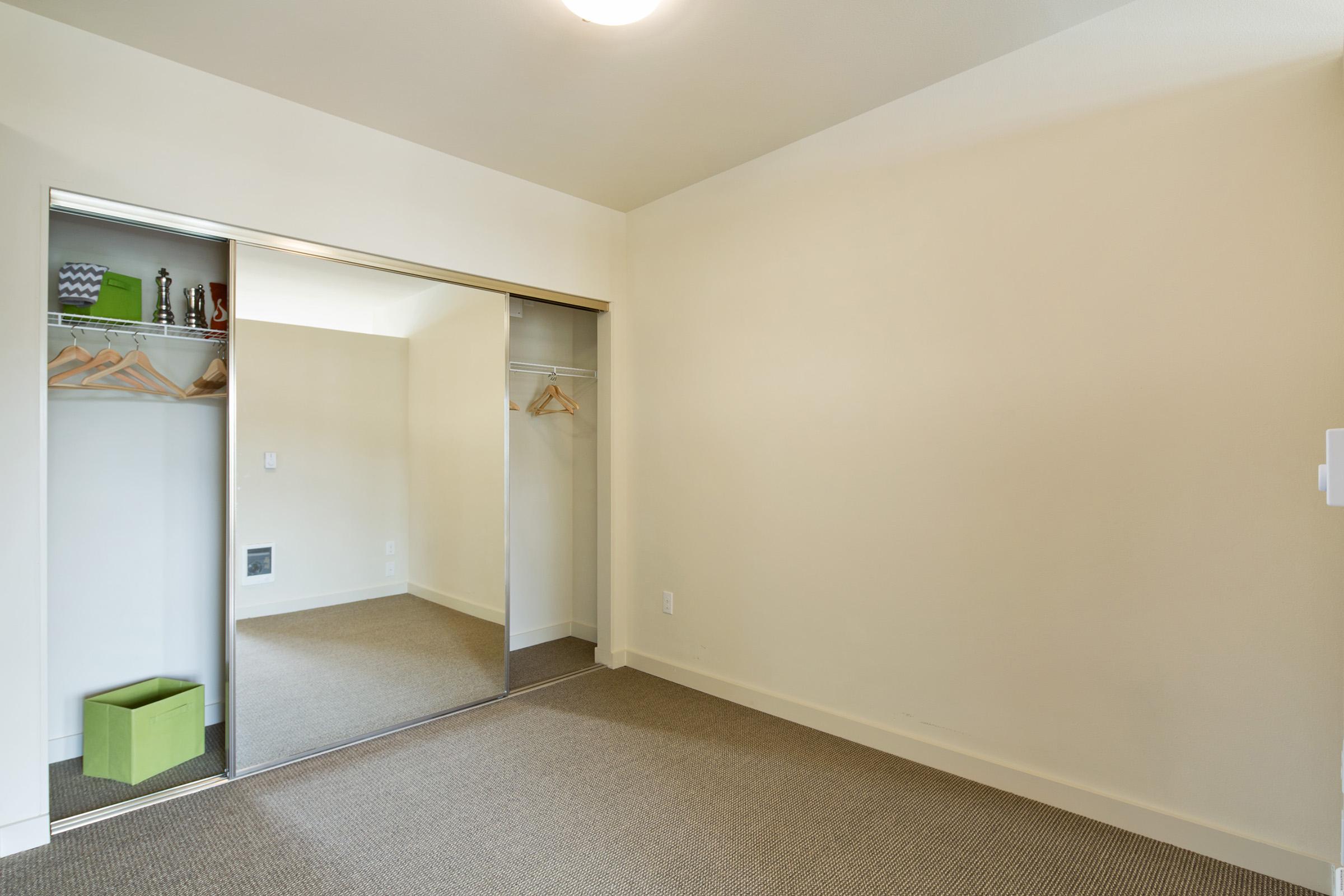 Empty room featuring a light beige wall and brown carpet. A mirrored sliding closet door is visible on the left, showcasing a few wooden hangers and a green storage box on the floor. T