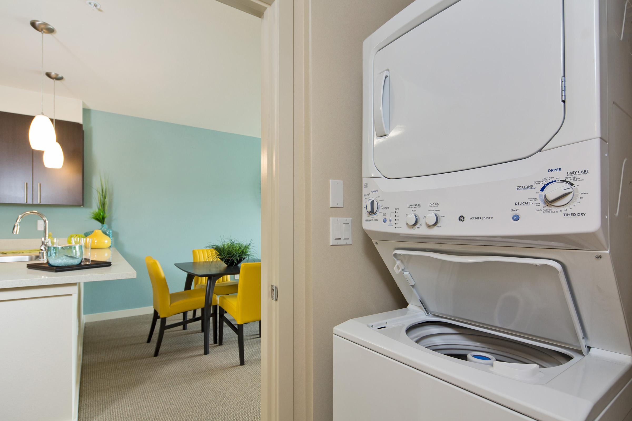 A stacked washer and dryer unit in a modern apartment setting, with a view of a kitchen that has dark cabinets and a small dining area featuring a round table and yellow chairs, all set against a light blue wall.