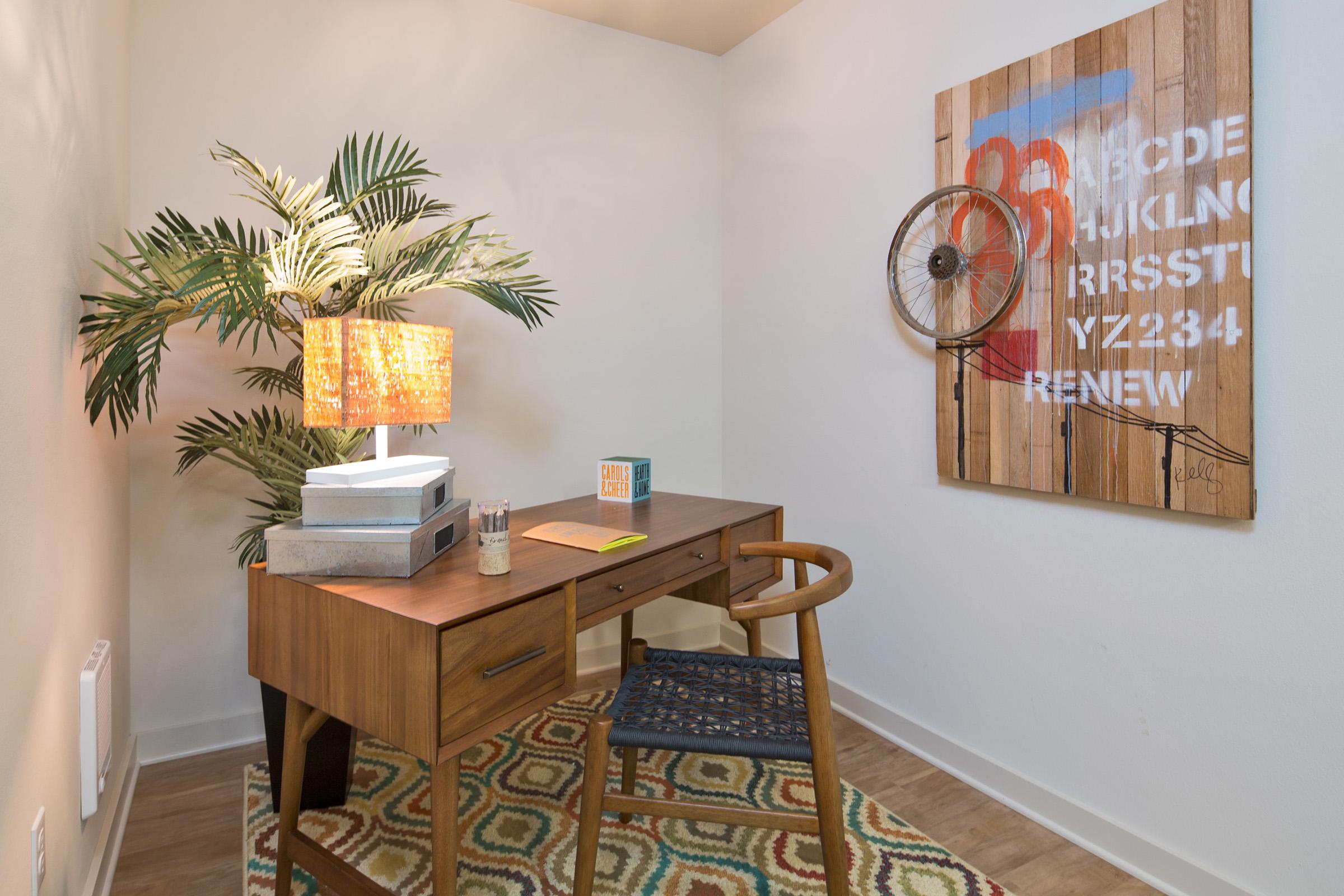 A small, stylish home office featuring a wooden desk with a chair, a lamp with a patterned shade, and decorative items.