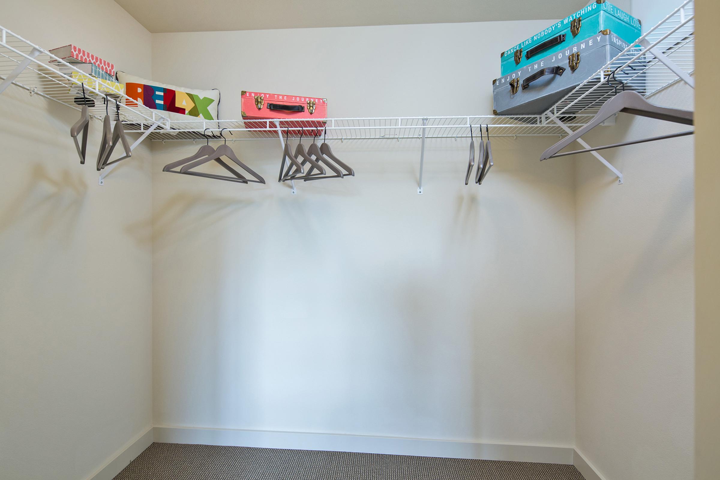 An empty closet featuring white wire shelves and several colorful suitcases. There are also multiple hangers hanging fr