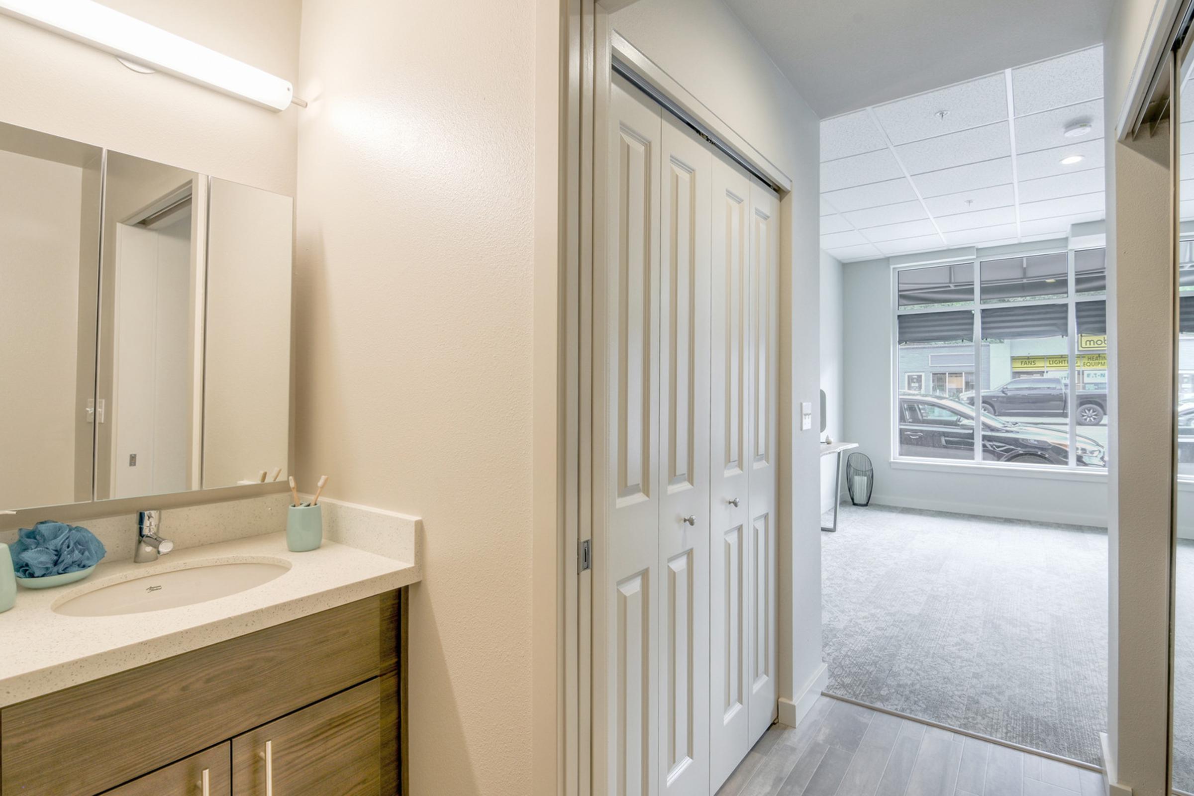 A well-lit bathroom with a modern vanity and a mirror, featuring a light-colored countertop and a small blue towel. There are two doors leading to other areas, and a spacious room visible through an open doorway, showcasing carpeted flooring and natural light from windows.