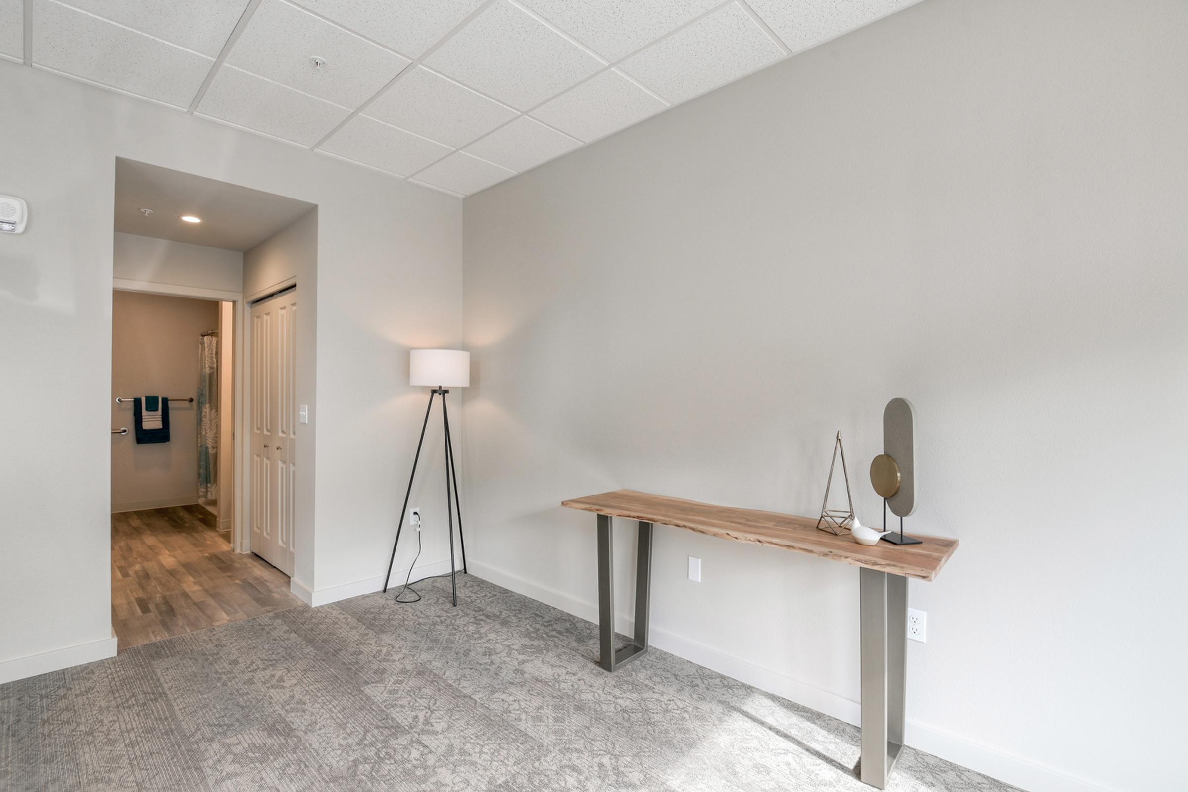 A minimalist room featuring a wooden table with decorative items, a standing lamp, and light gray walls. A doorway leads to a bathroom area, while the floor is covered with neutral-colored carpet. The space has a modern and airy feel, with soft lighting and simple decor.