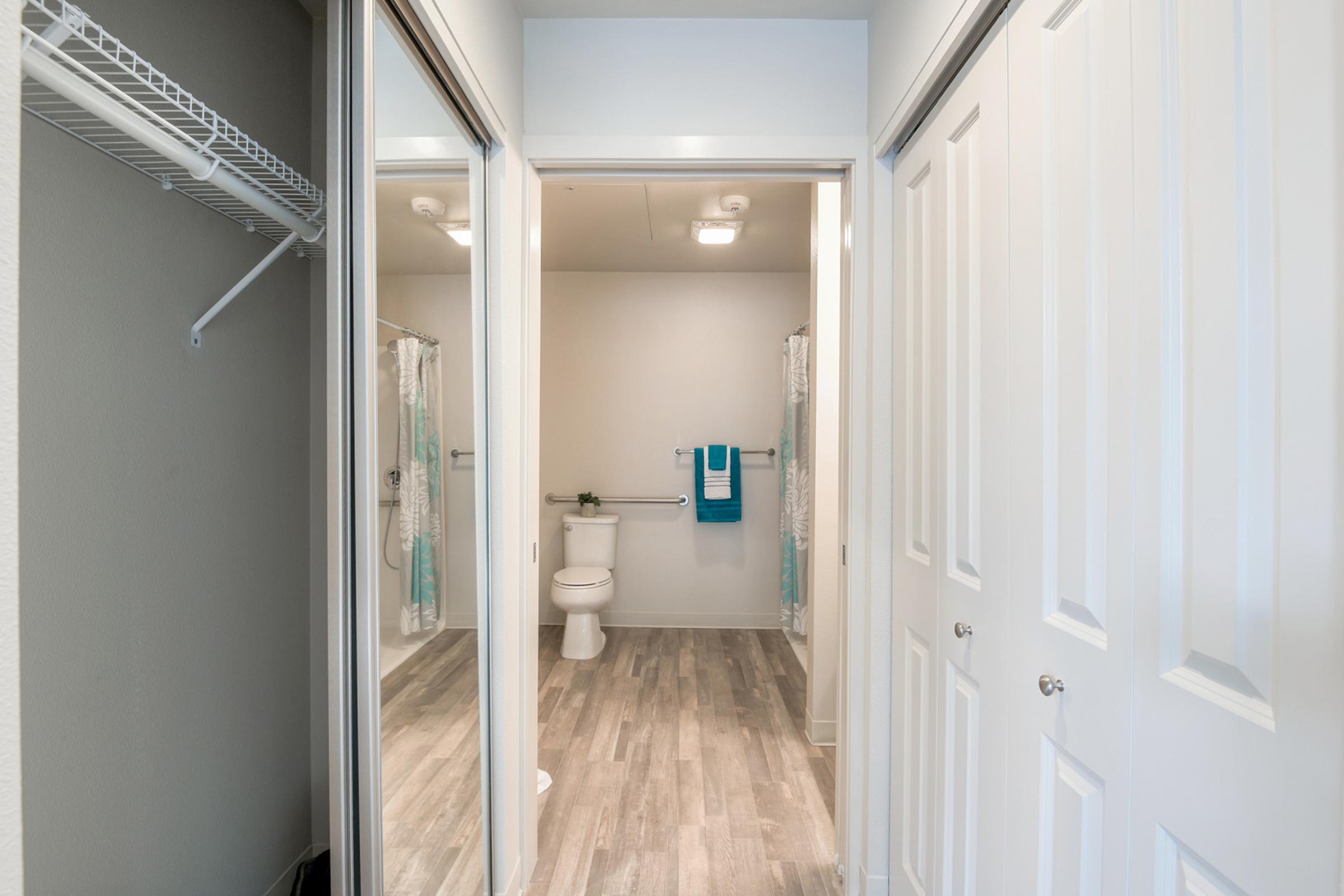 A well-lit bathroom with a shower, toilet, and towels hanging on a towel rack. The floor is covered in light wood-like tiles, and there are two white closet doors visible to the left. The walls are painted in a light color, enhancing the spacious feel of the area.