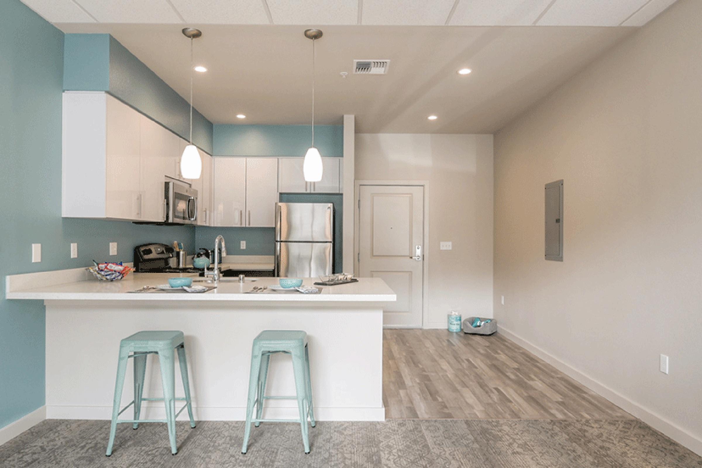 A modern kitchen with light blue accent walls, featuring white cabinets and stainless steel appliances. A central island with two turquoise stools offers seating. The space is well-lit with pendant lights and has a neutral-colored floor, creating a clean and inviting atmosphere.