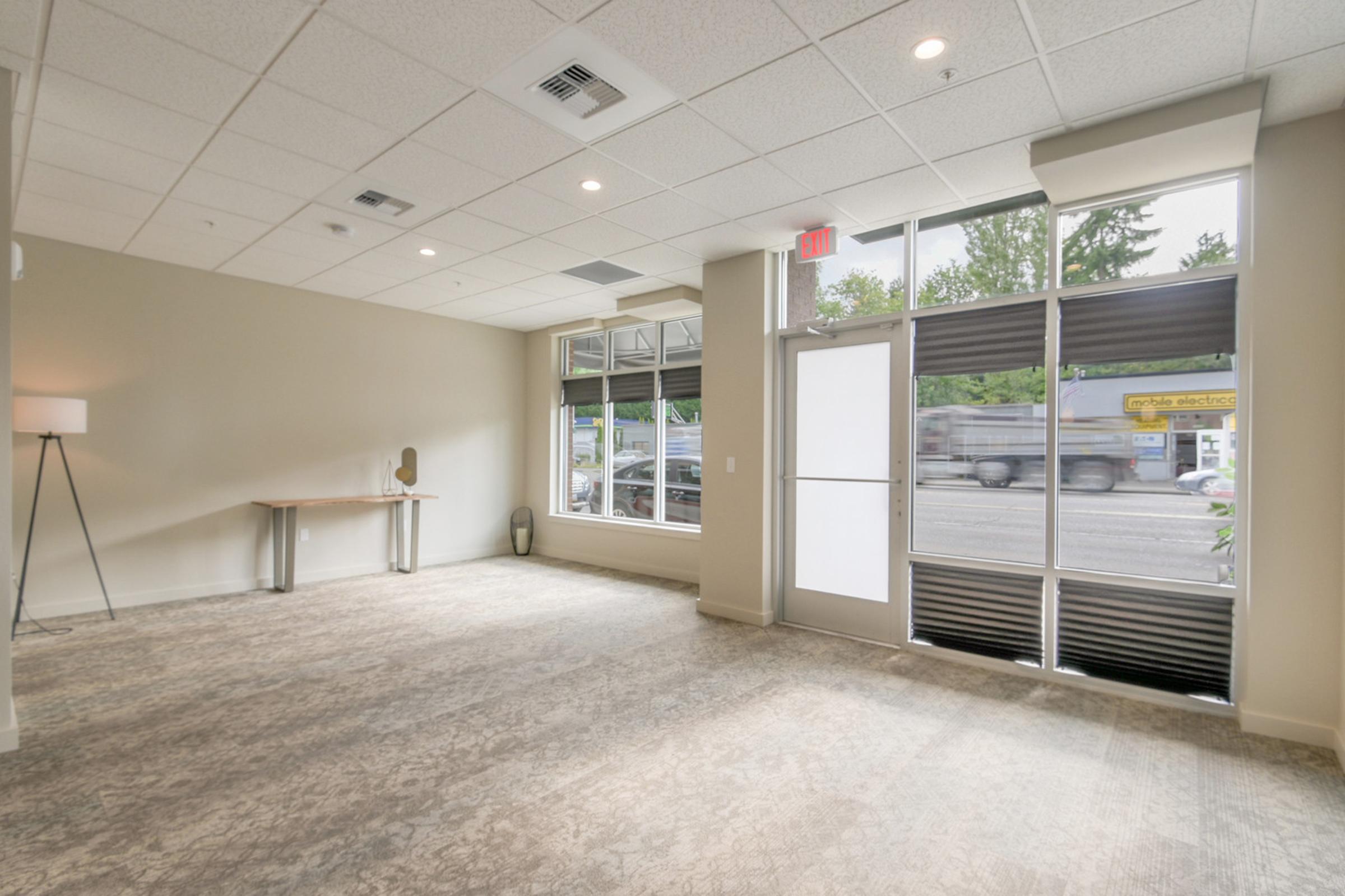 A spacious, well-lit interior of a commercial space with beige walls and carpet. Large windows provide natural light, and there's a minimalistic table and a lamp. The entrance features glass doors leading to a sidewalk with street views. The overall ambiance is modern and inviting.