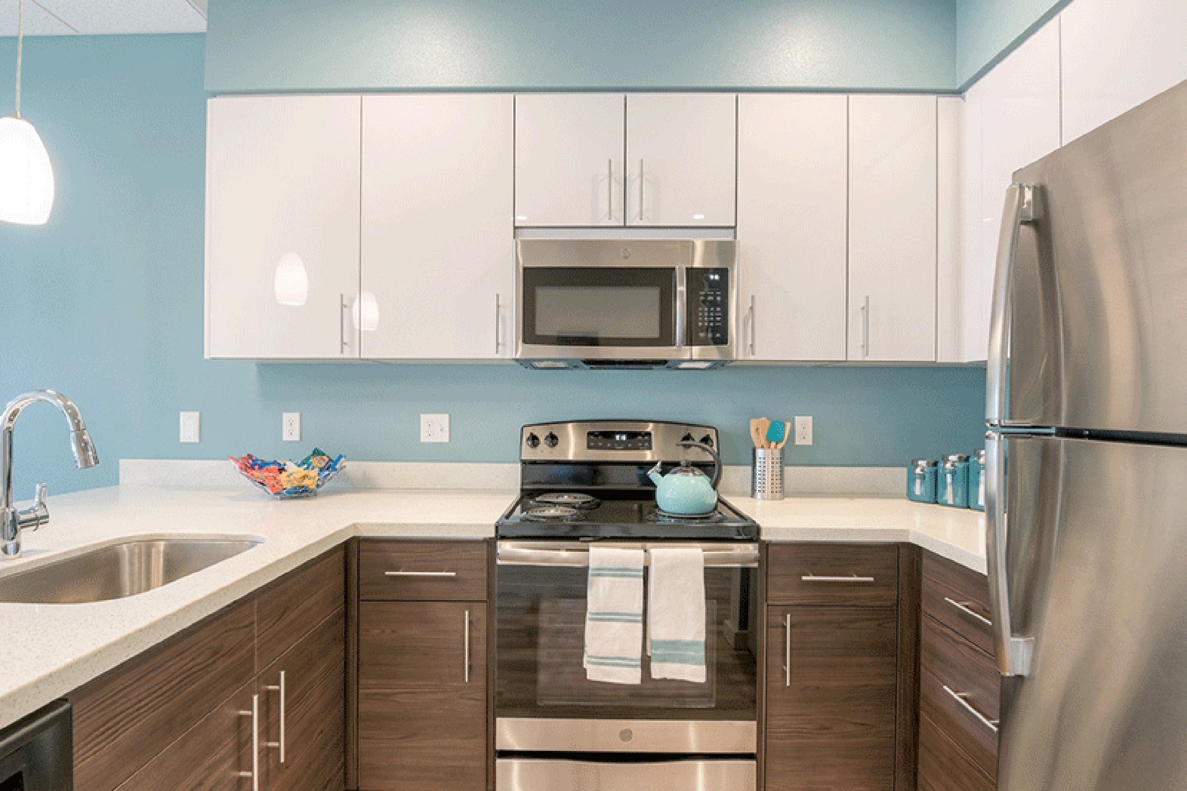 Modern kitchen featuring a stainless steel stove, microwave above, and a double sink. The cabinetry is a mix of white and dark wood, with a light blue wall accent. A turquoise teapot is on the stove, and colorful dishware is displayed in a bowl on the counter. Bright and inviting ambiance with pendant lighting.
