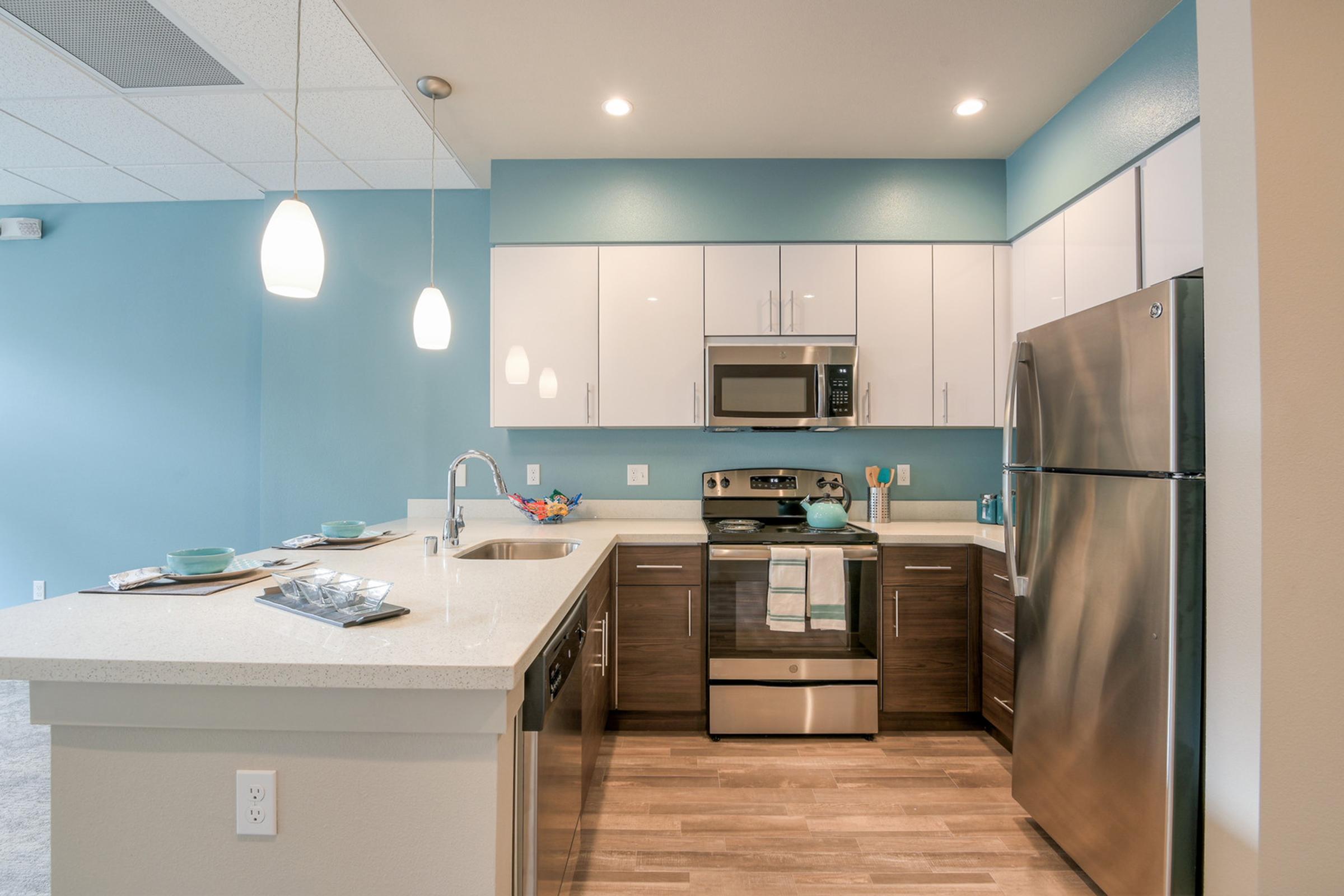 Modern kitchen with light blue walls, white cabinets, and stainless steel appliances, including a refrigerator and microwave. Features a countertop with a sink, a small dining area with teal plates, and pendant lighting. The flooring is light wood, creating a bright and inviting atmosphere.