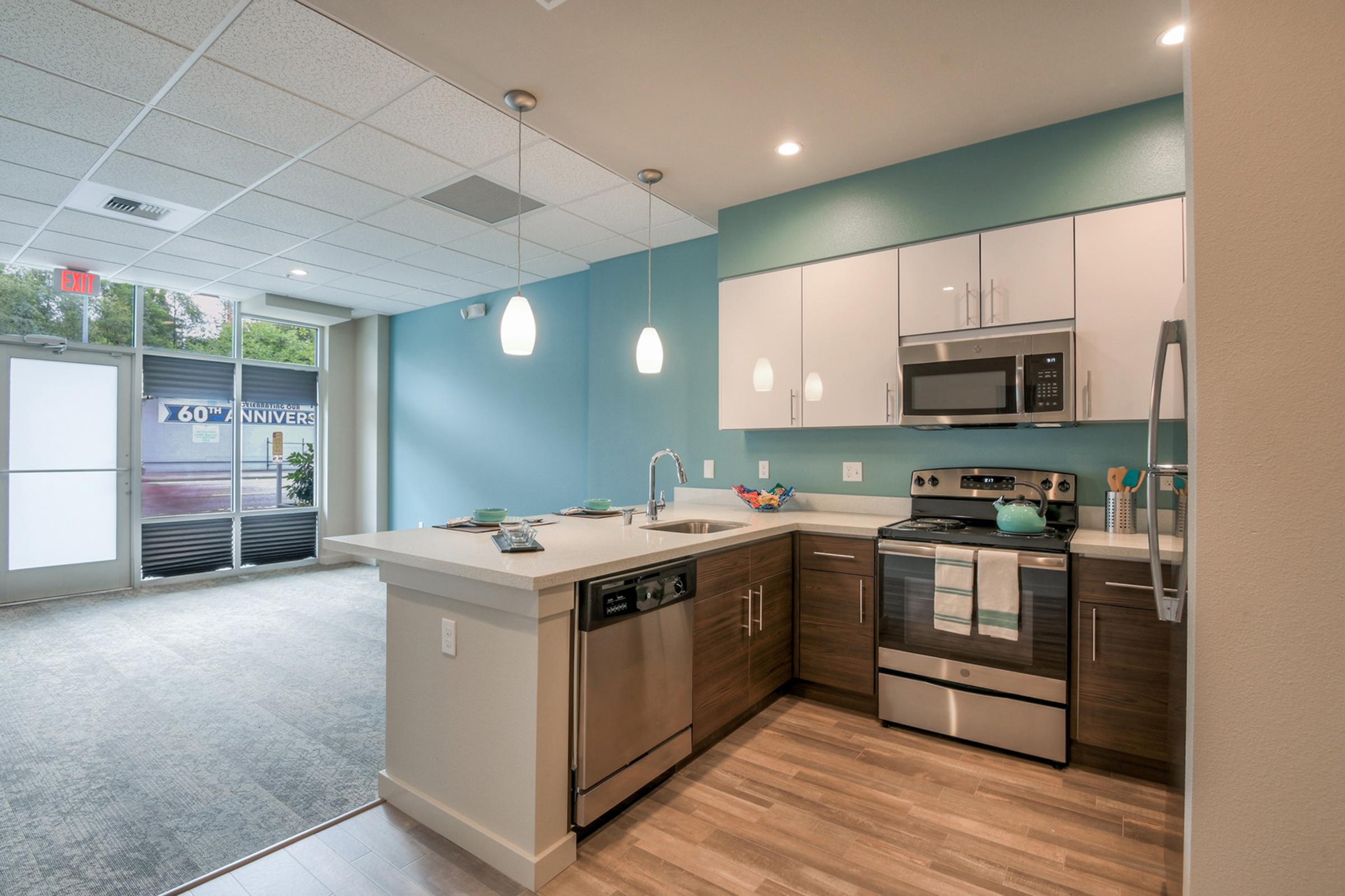 Modern kitchen with a combination of light blue and white cabinetry. Features include stainless steel appliances, a stainless steel sink, and a dishwasher. A large countertop provides ample workspace. The space is well-lit with pendant lights and has an open layout leading to a door labeled '60’ UNIVERSE.'