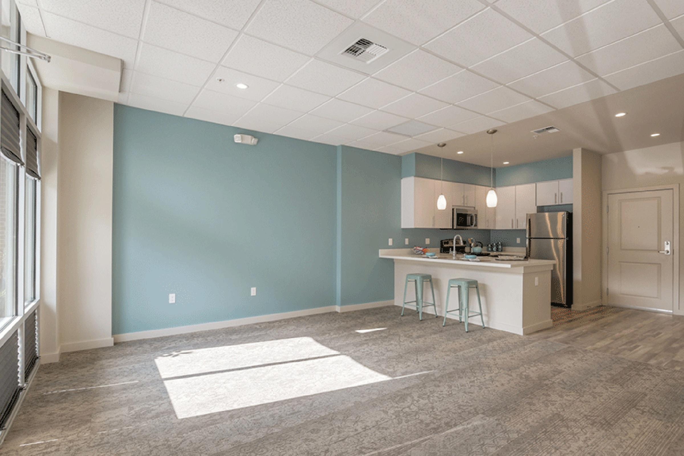 Spacious modern living area with light blue walls, featuring a kitchen with stainless steel appliances, white cabinetry, and bar stools. Large windows allow natural light to fill the room, highlighting the plush carpeted floor.