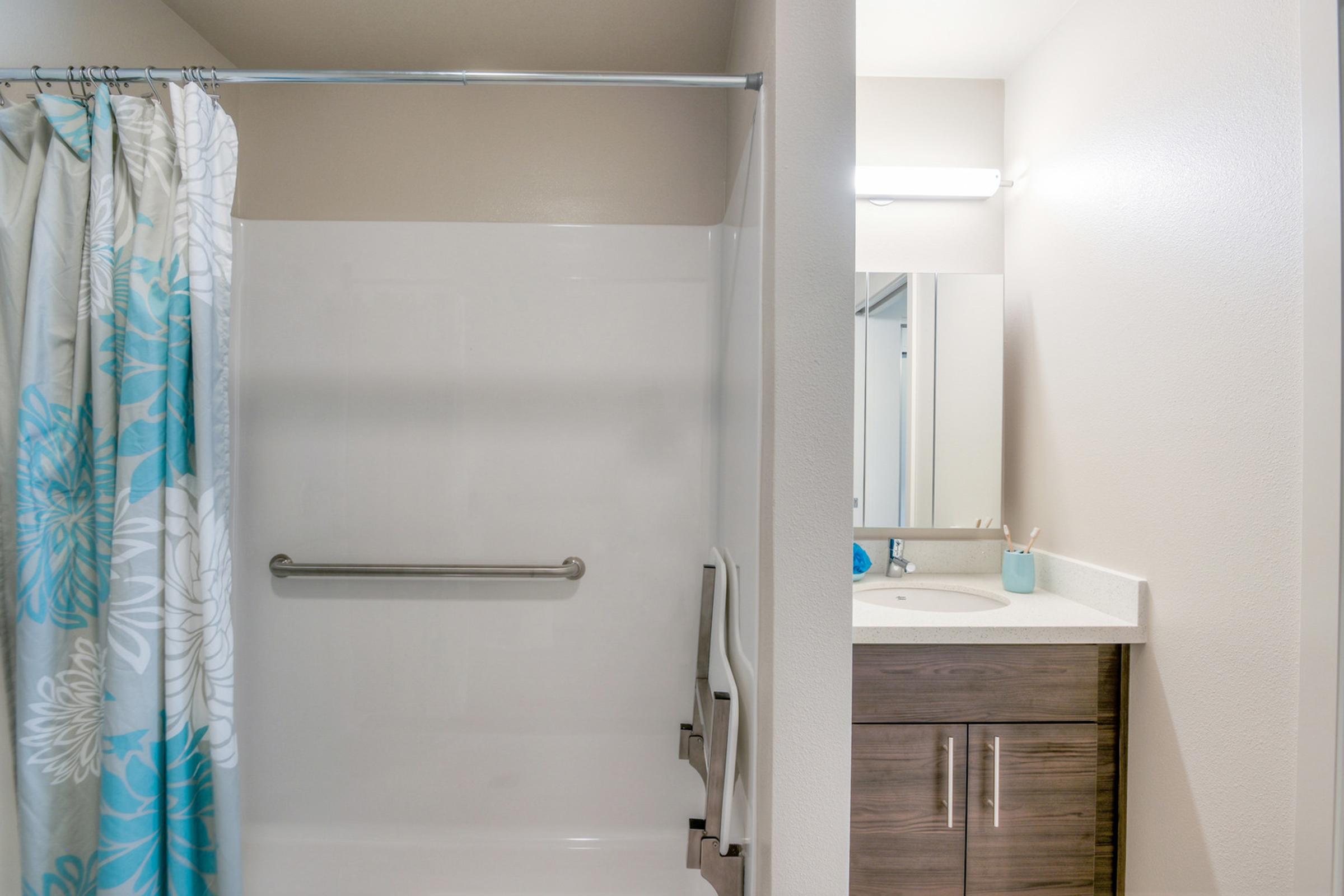 A bright and clean bathroom featuring a shower with a blue and white floral curtain. To the right, there is a vanity with a sink and a modern mirror, accompanied by a small blue container and toothbrushes. The walls are painted in a light color, enhancing the spacious feel of the room.