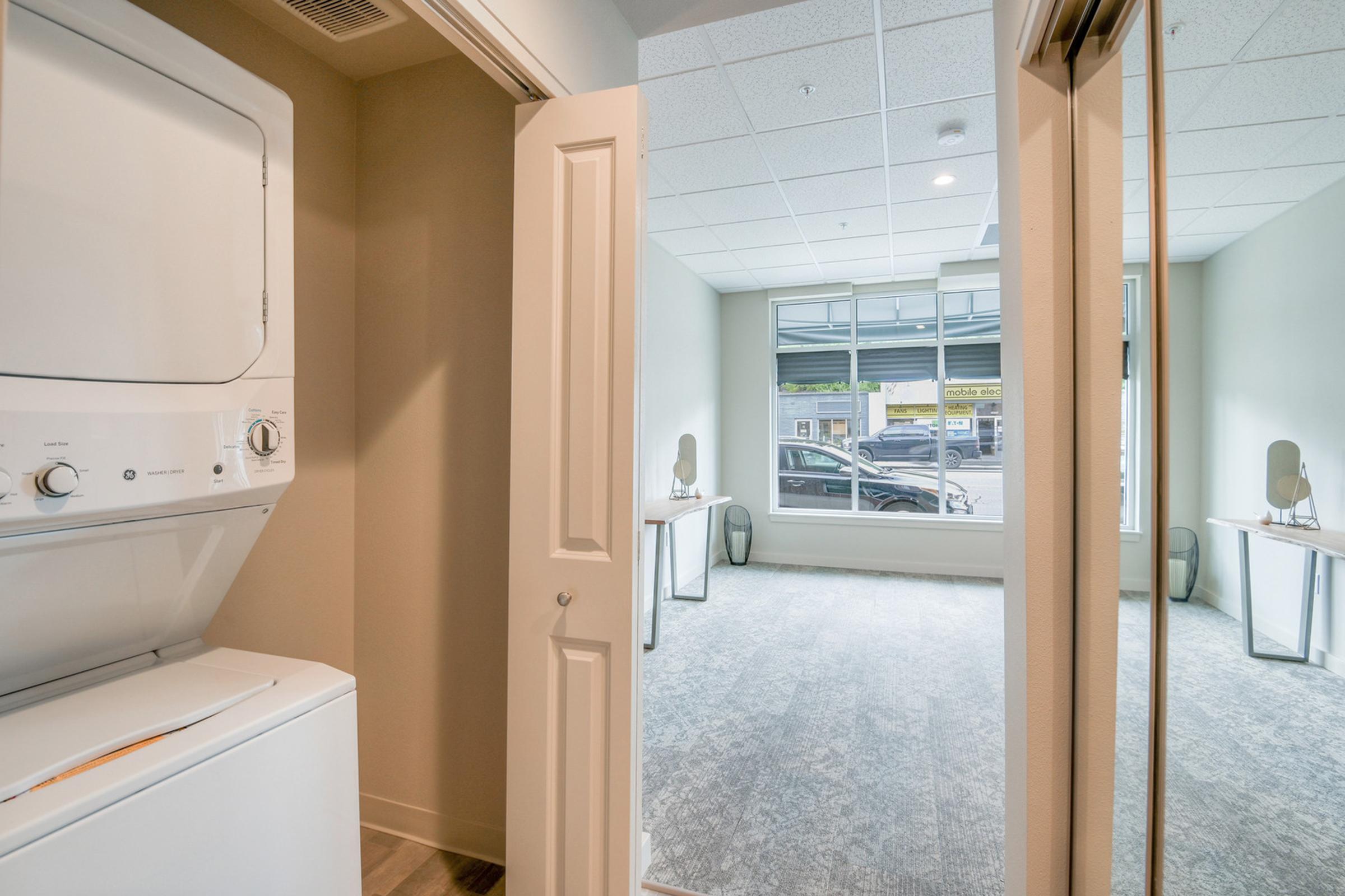 View of a laundry area with a stacked washer and dryer on the left. To the right, a spacious room with large windows, grey carpet, and two tables with decorative items. Natural light fills the room, creating a bright and open atmosphere.