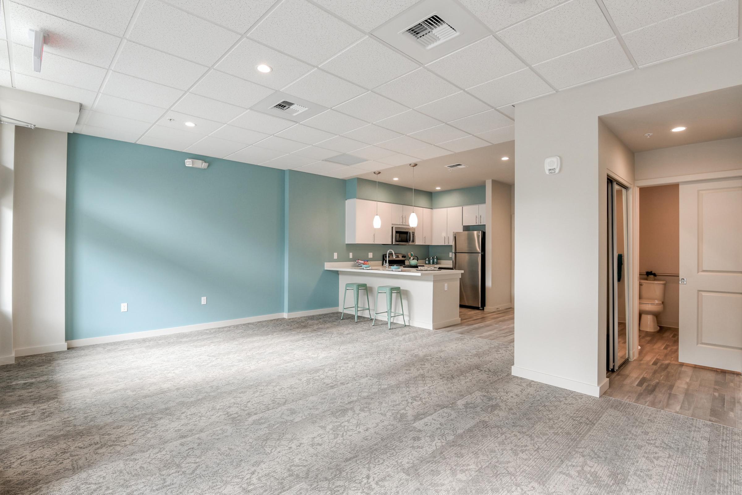 A spacious, modern apartment interior featuring a light blue accent wall, an open layout with a kitchen bar area, and sleek appliances. The flooring is light, and large windows allow natural light to fill the room. A bathroom entrance is visible in the background.