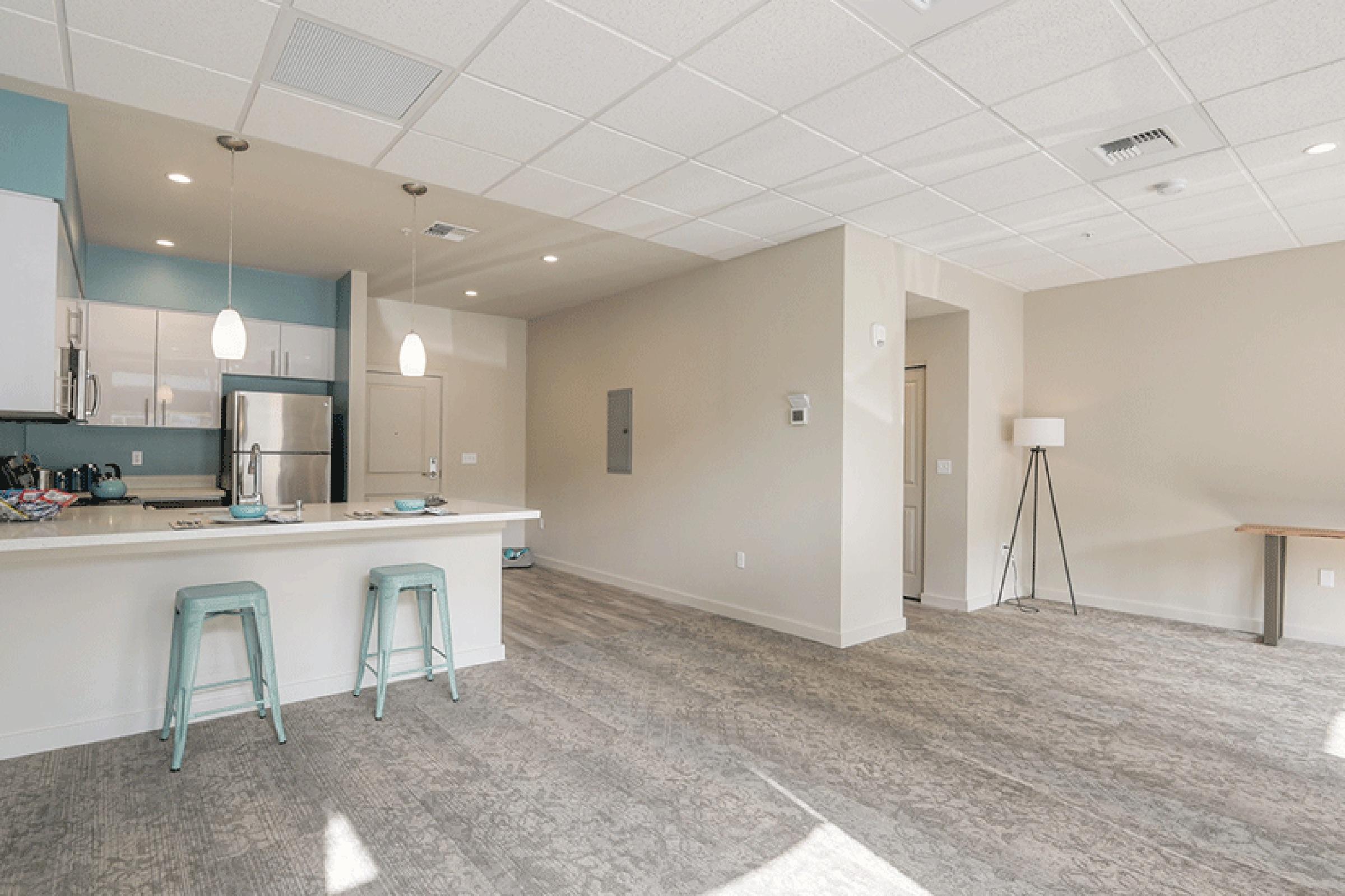 A modern, open-concept apartment kitchen and living area. The kitchen features white cabinets, stainless steel appliances, and light blue accents. A breakfast bar with turquoise stools is visible. The living space is empty with large windows allowing natural light, and a minimalist floor lamp stands in the corner.