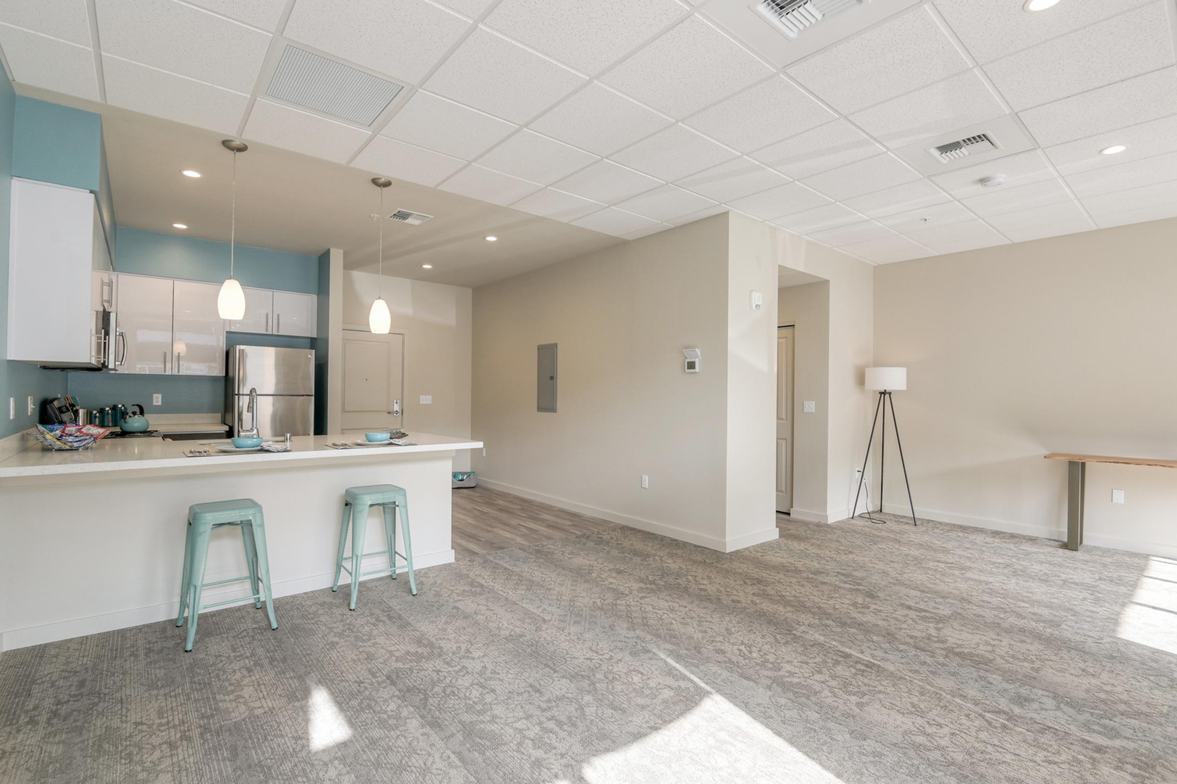 Modern open-concept living space featuring a kitchen with light blue cabinetry and stainless steel appliances. A dining area with two teal bar stools is adjacent to the kitchen. The flooring is light gray, with ample natural light illuminating the room. A minimalist lamp and a simple table are positioned in the background.