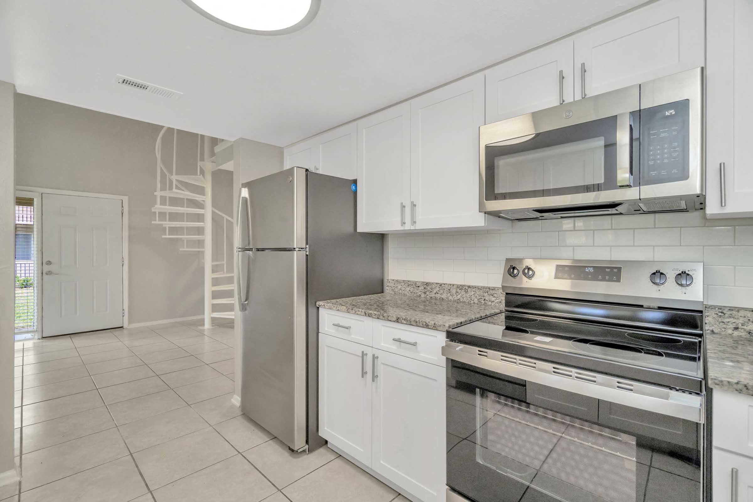 A modern kitchen featuring stainless steel appliances, including a microwave and oven, with a granite countertop. There is a silver refrigerator and white cabinets above the counter. The background shows a staircase leading to a second level, and the flooring is tiled in a neutral color.