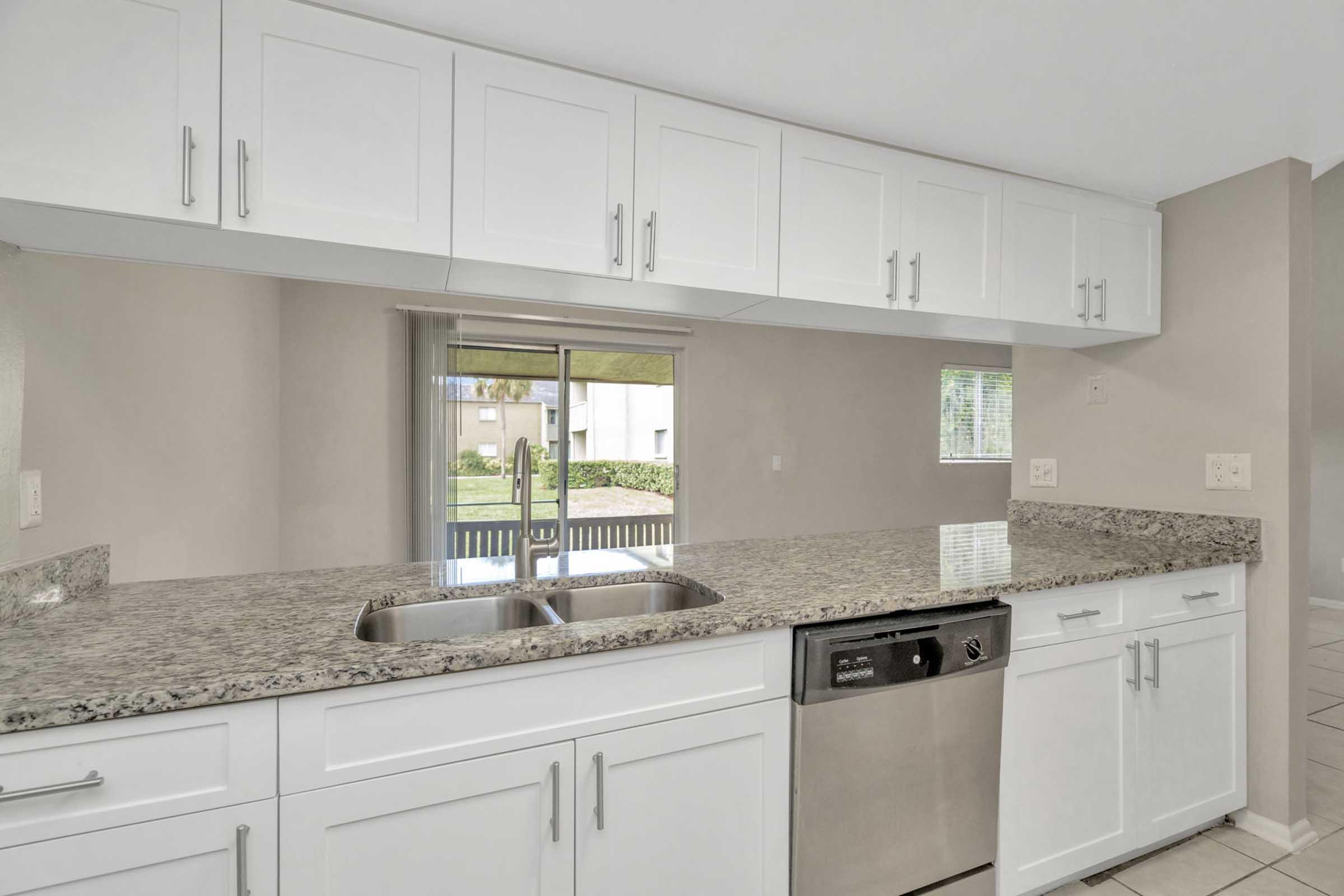 A modern kitchen featuring white cabinets and a granite countertop with a double sink. A stainless steel dishwasher is visible beneath the counter. Large windows provide natural light, and there's a view of an outdoor space beyond. The floor is tiled, and the overall color scheme is light and airy.