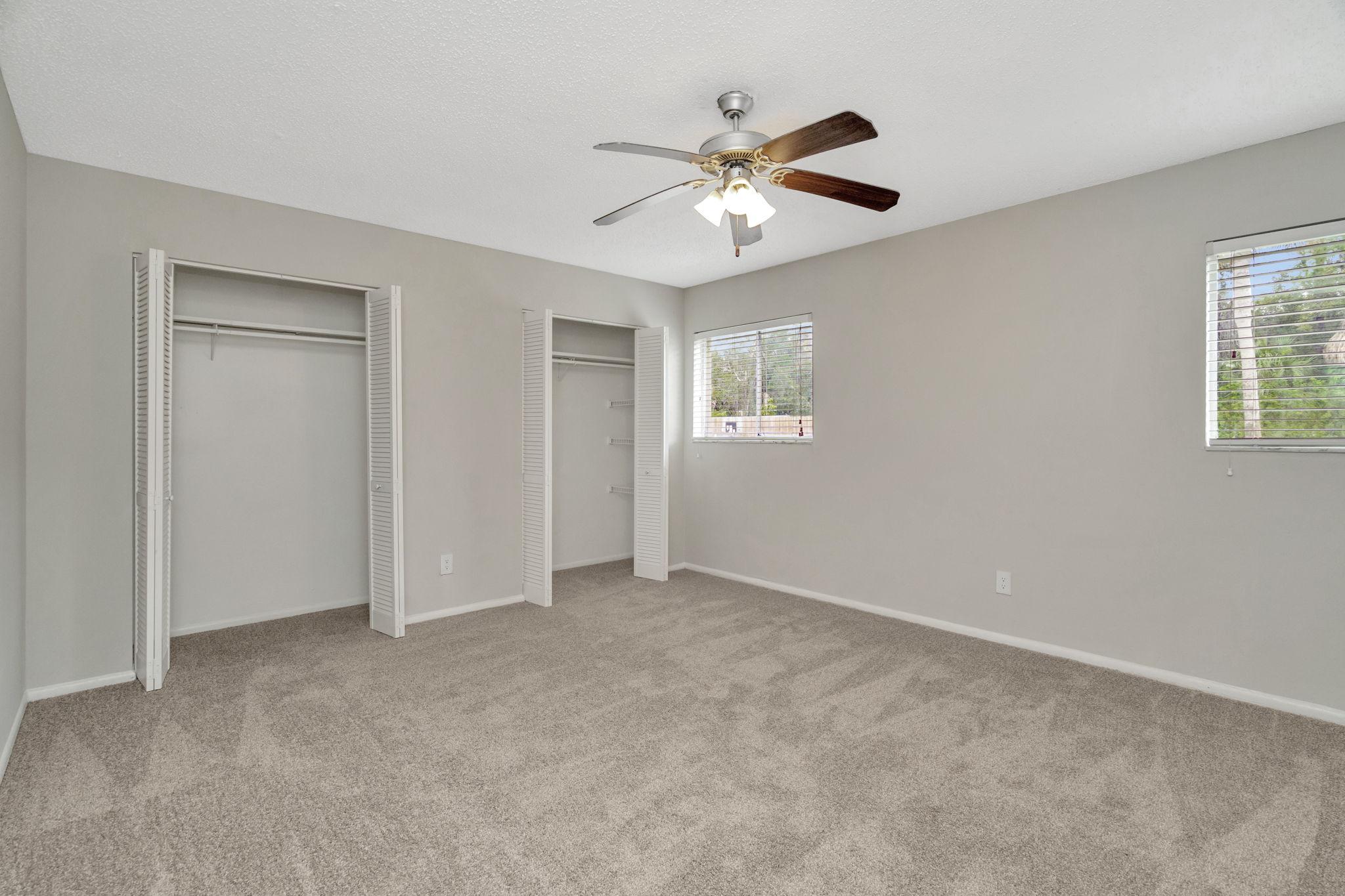Spacious empty bedroom with freshly carpeted flooring, a ceiling fan, light gray walls, and two windows providing natural light. It features two closets with sliding doors, ready for personalization and furnishing.
