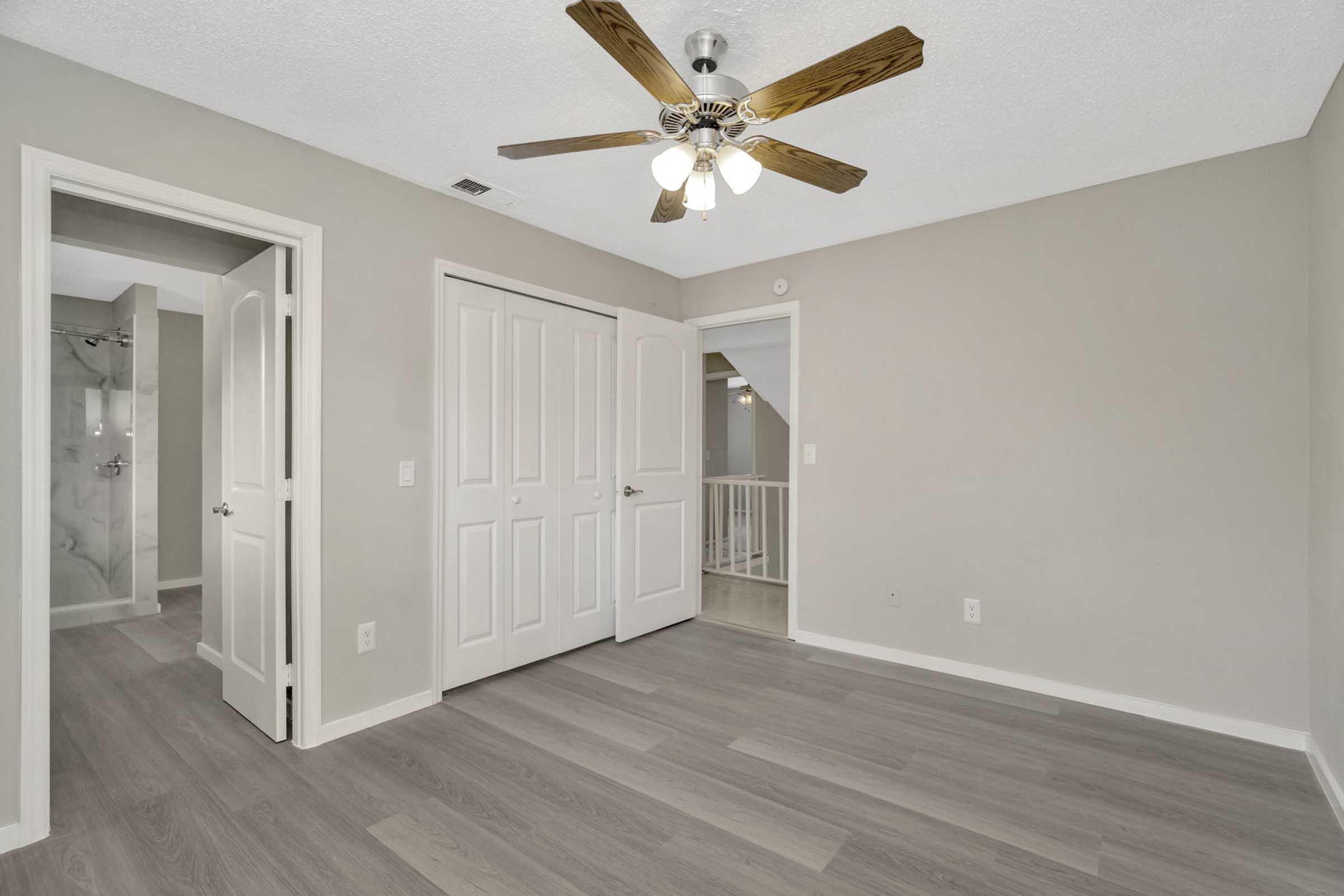 A well-lit room featuring light gray walls and modern wood flooring. There is a ceiling fan with four lights, double doors leading to a closet, and an open door that provides access to a bathroom or another room. The decor is minimalist and contemporary, creating a spacious atmosphere.