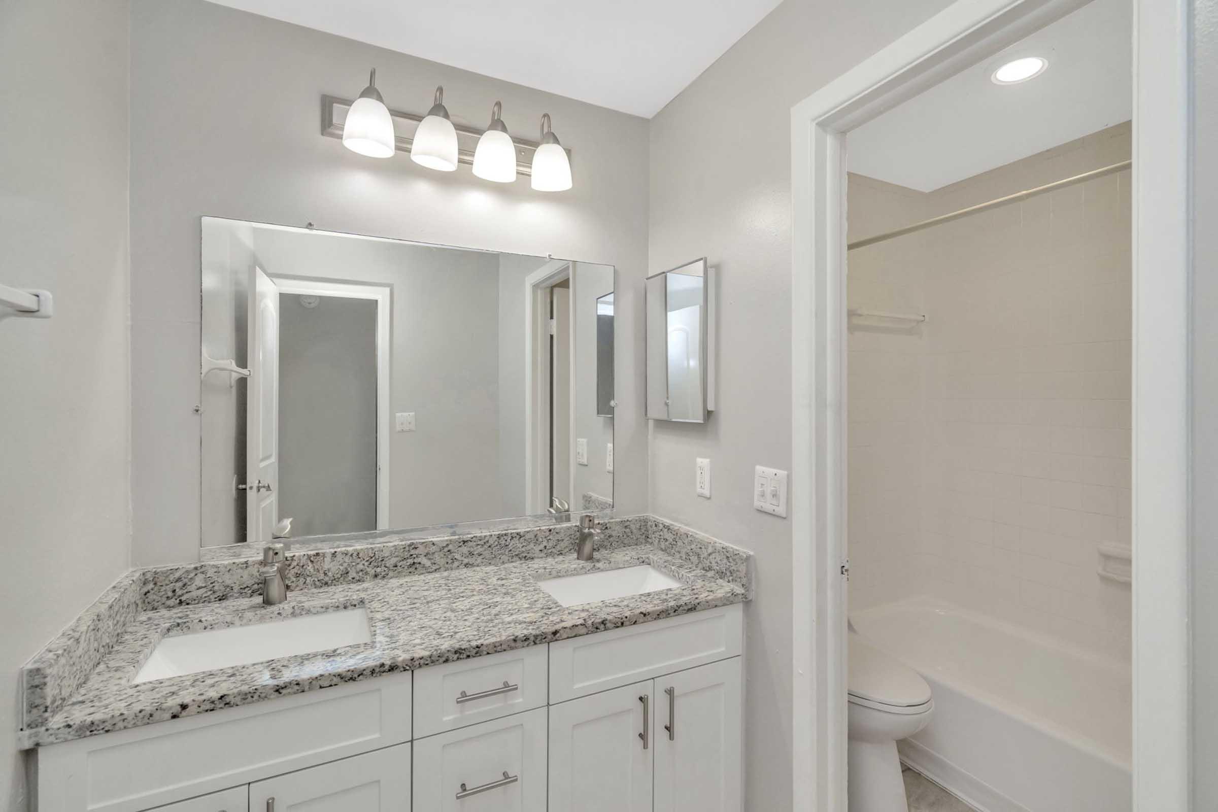Modern bathroom with a double sink vanity featuring a speckled granite countertop, two mirrors, and stylish lighting fixtures. The space has light-colored walls and a clean, minimalistic design, along with a bathtub and white tiles in the shower area. The overall look is bright and inviting.