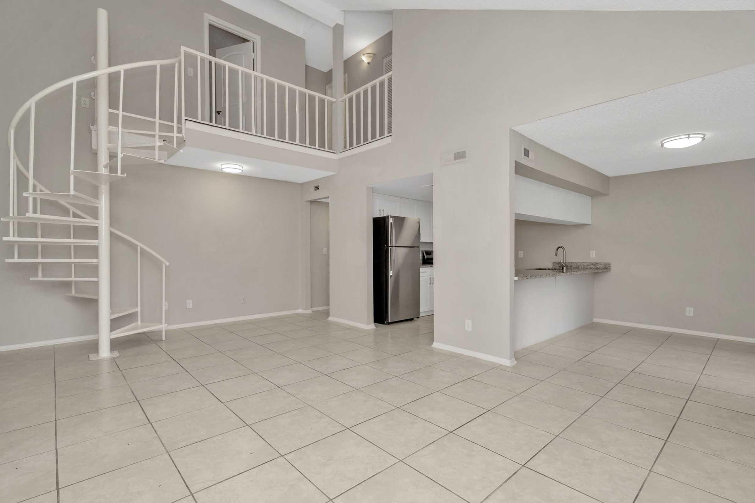A spacious open-concept living area featuring light-colored walls, a tiled floor, and a spiral staircase. To the right, there is a kitchen area with a bar counter, and a stainless steel refrigerator is visible. The space is well-lit with natural light coming from large windows above.