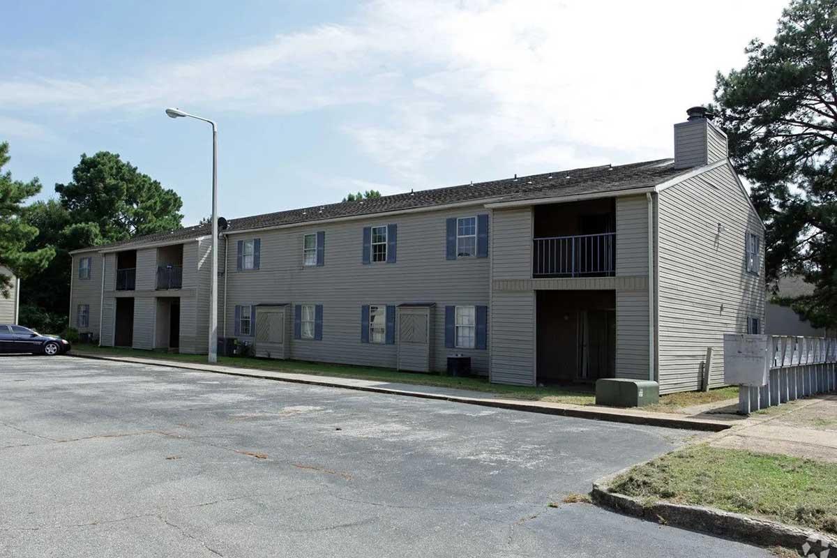 A two-story apartment building with light gray siding, featuring several windows and balconies. The building is situated beside a paved parking lot, with a few trees in the background and a streetlamp nearby. The surrounding area appears well-maintained and residential.