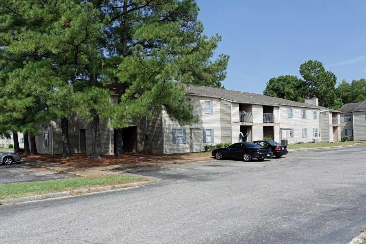 A multi-unit residential building surrounded by tall pine trees. The building has multiple windows and balconies, with a parking lot in front that features several parked cars. The scene is set on a clear day with blue sky and greenery in the background.