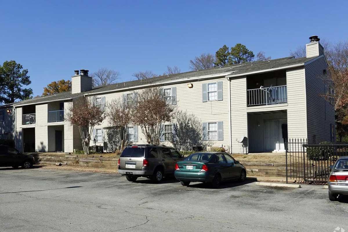 Two-story apartment building with multiple units, featuring a mix of balconies and ground-level entrances. Several parked cars are in the foreground, and the surrounding area includes sparse trees and grass. The sky is clear and blue, indicating a bright day.