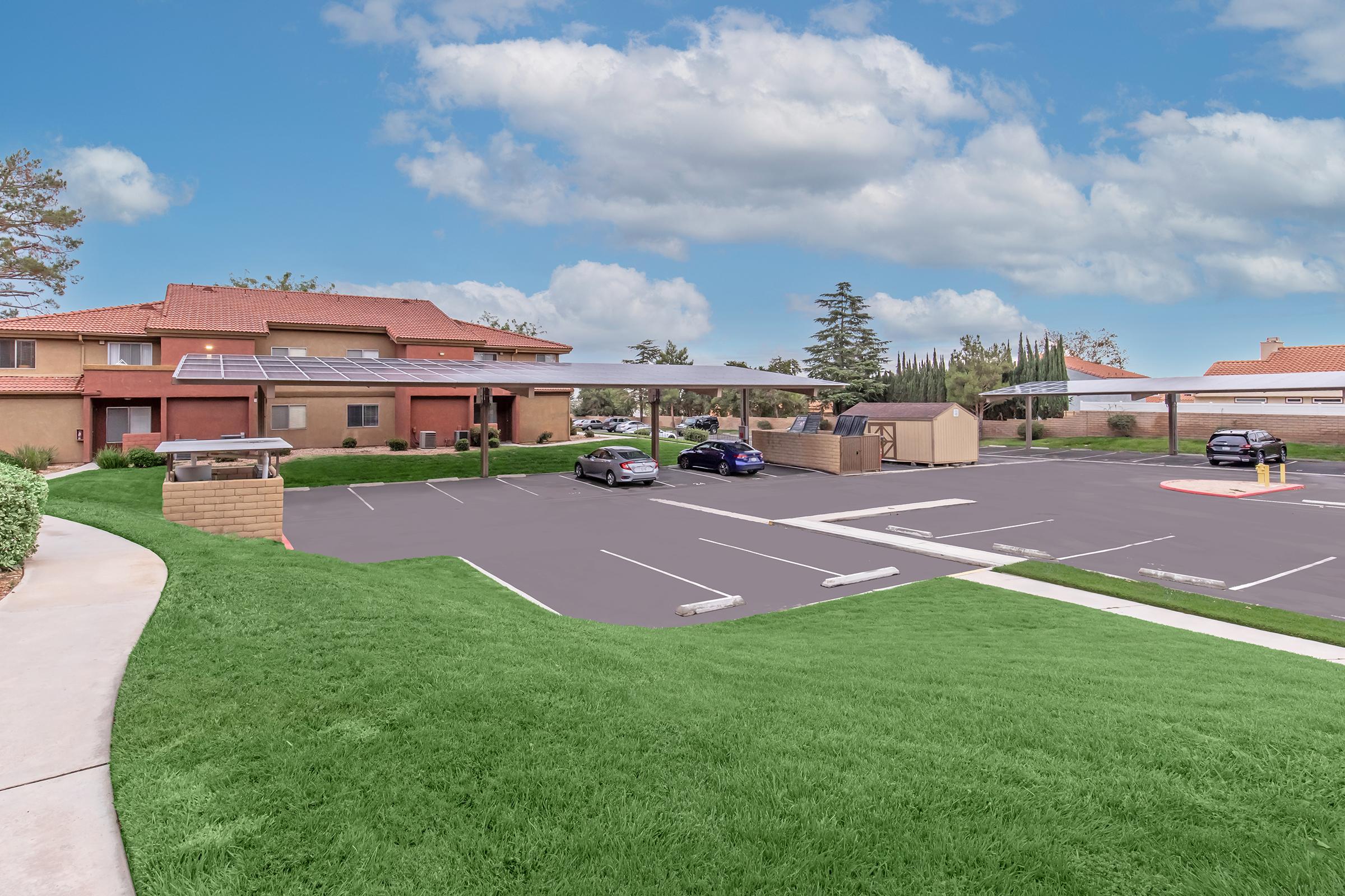 A residential complex with two buildings featuring solar panels on the roofs. In the foreground, a landscaped grassy area leads to a parking lot with several parked cars under shaded canopies. Clear blue sky with fluffy clouds overhead.